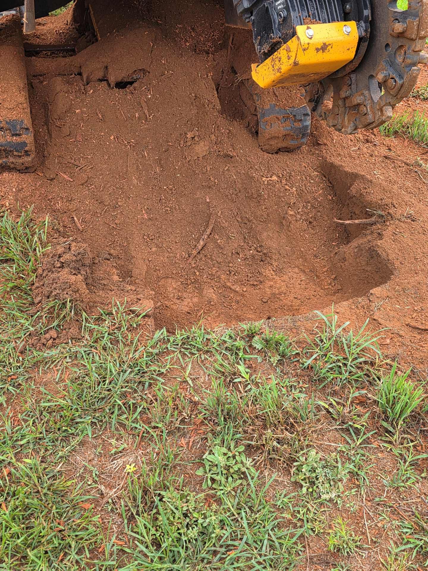 Yellow excavator bucket dumping red soil onto grass and dirt ground
