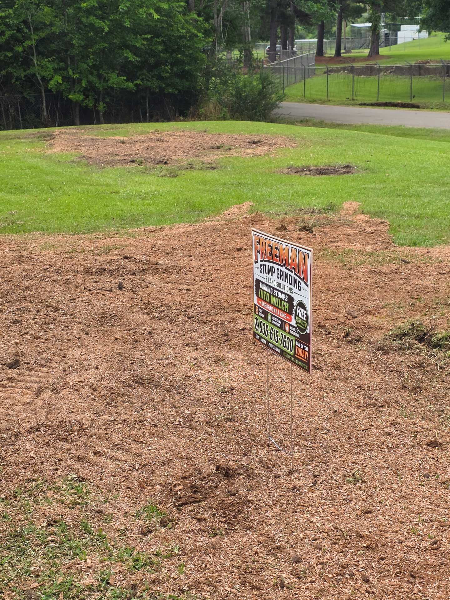 Mulched garden plot with a small sign in a grassy park near a fence and trees