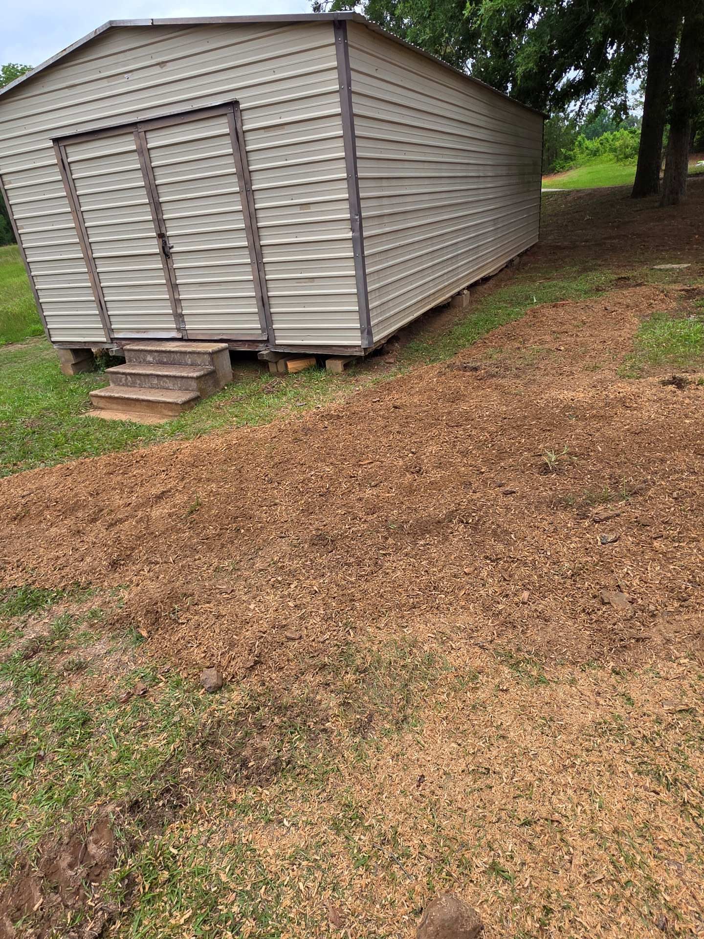 Beige storage shed with double doors on a gravel and dirt yard beside trees