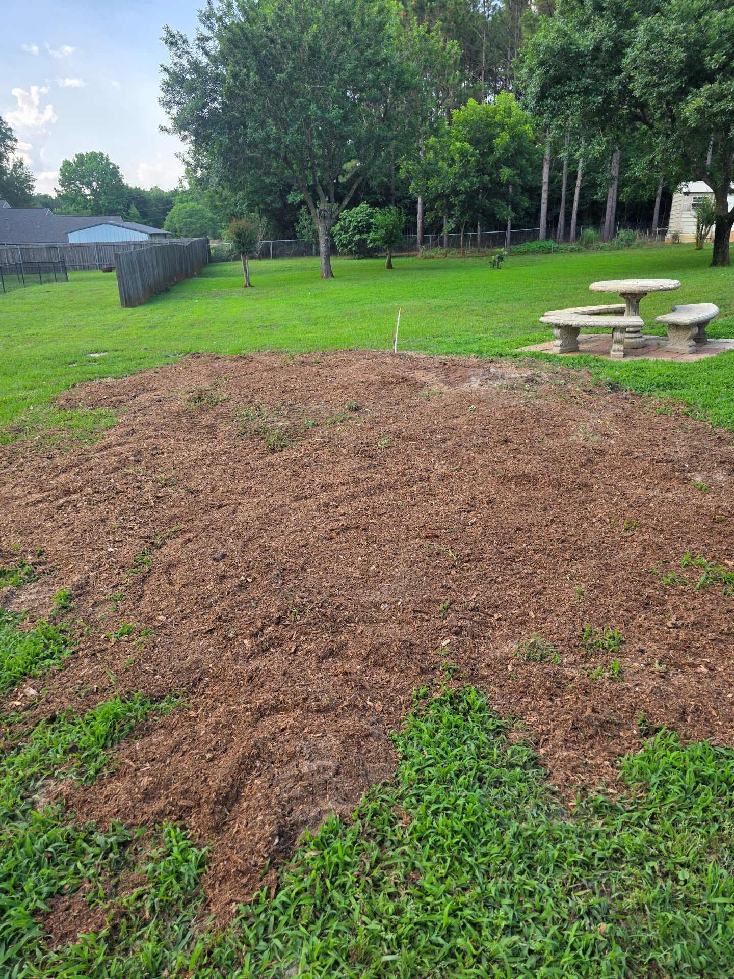 Freshly mulched garden bed in a grassy park with picnic tables and trees in the background