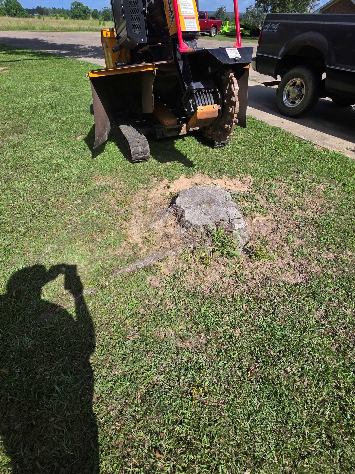 Backhoe beside a tree stump in a grassy yard, with a pickup truck parked nearby.