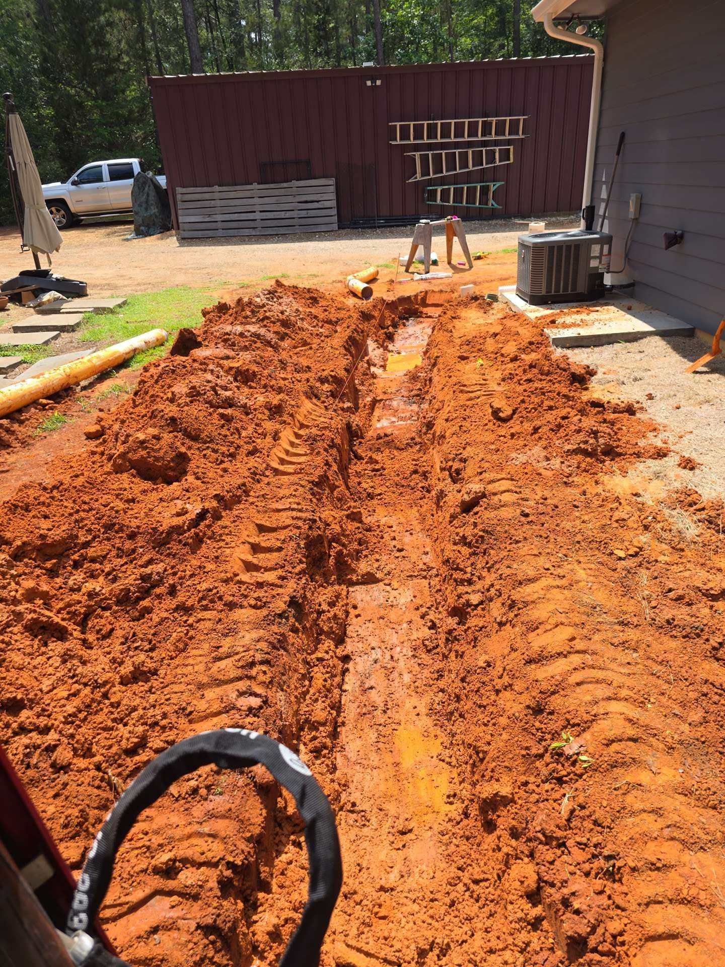 Freshly dug trench of orange dirt beside a house, with a bucket in the foreground and a work truck in back