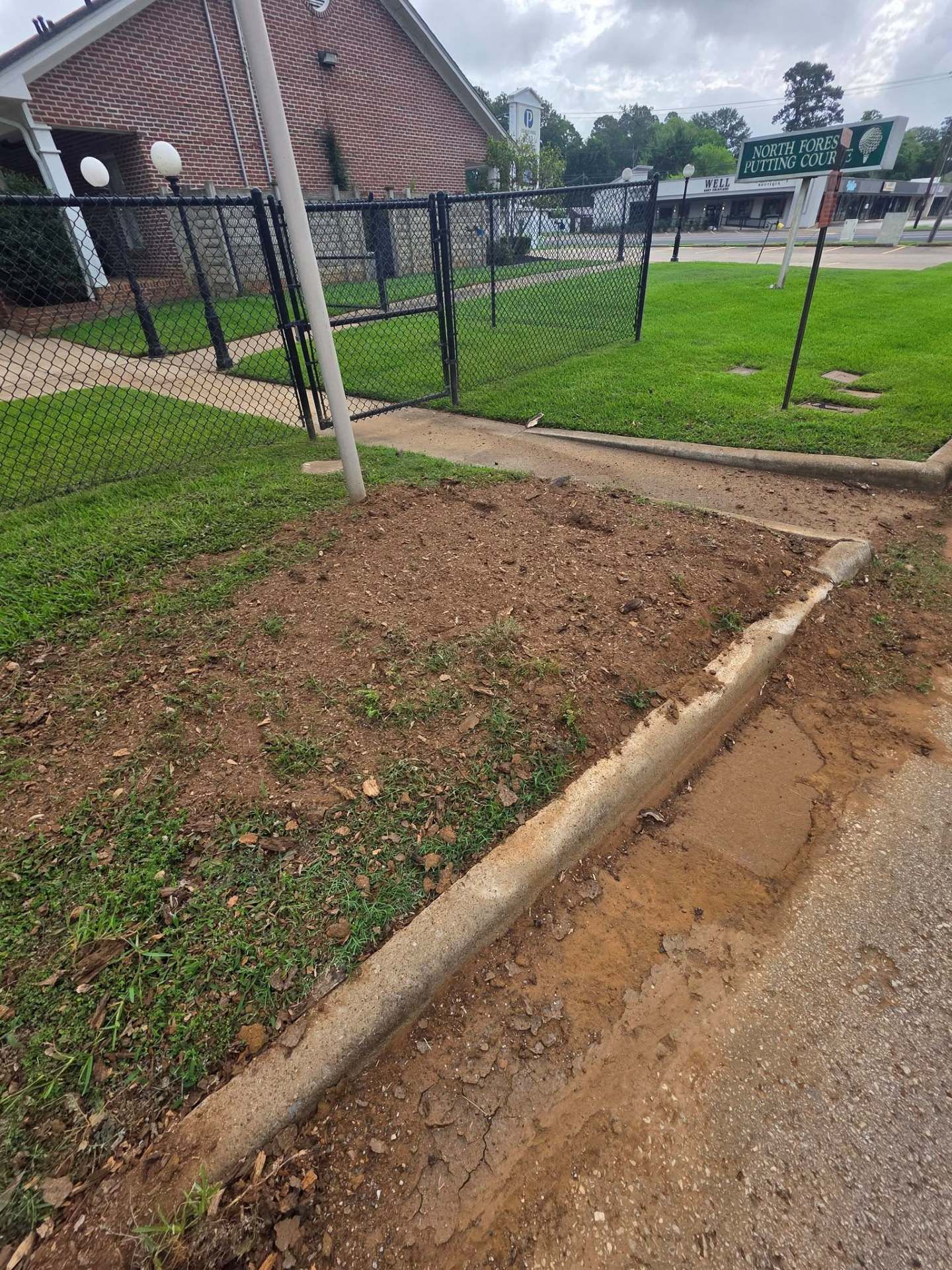 Small dirt patch beside a concrete sidewalk and grassy yard near a brick building and black fence