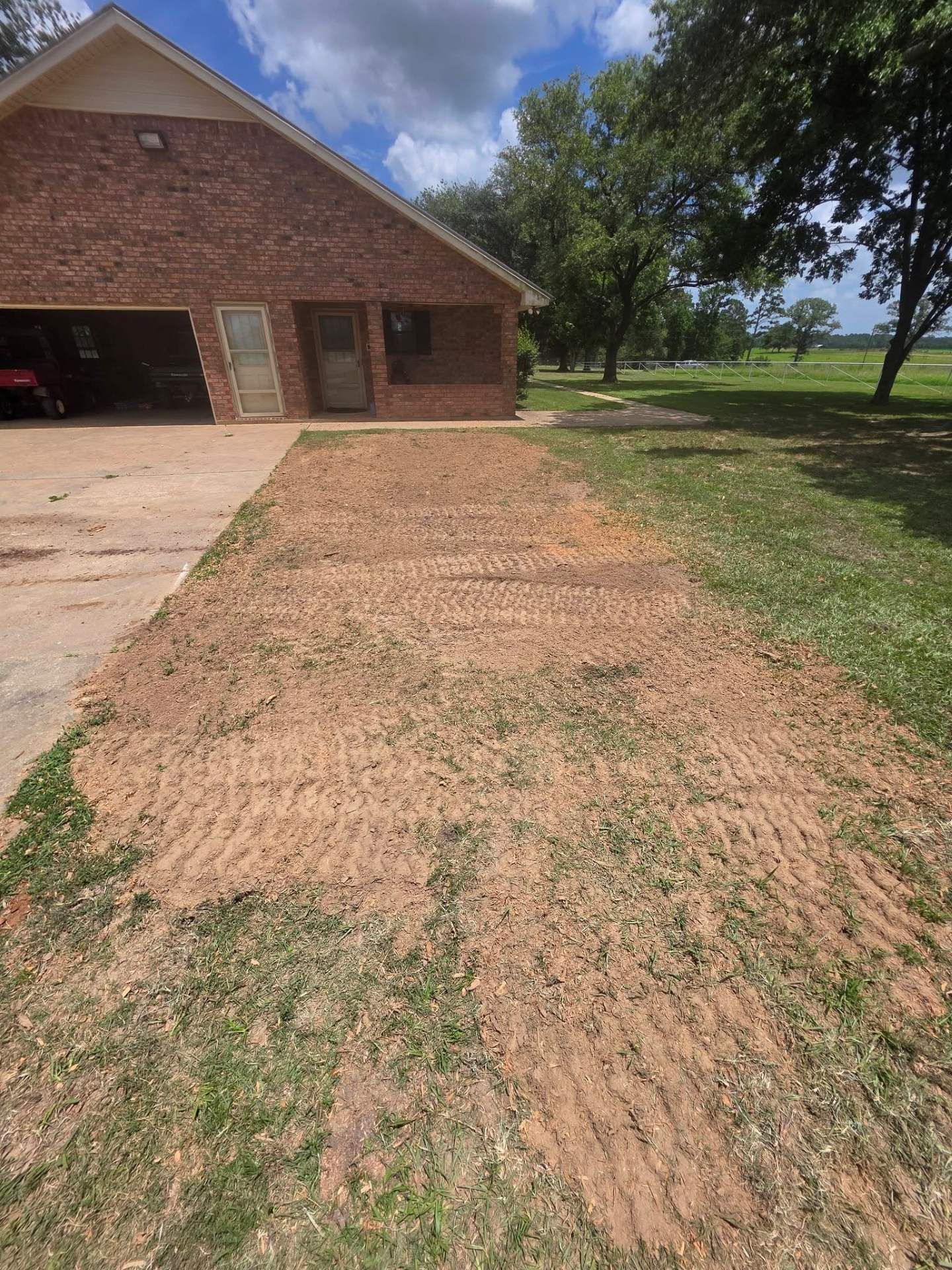 Brick house with a driveway, muddy yard, and trees under a partly cloudy sky