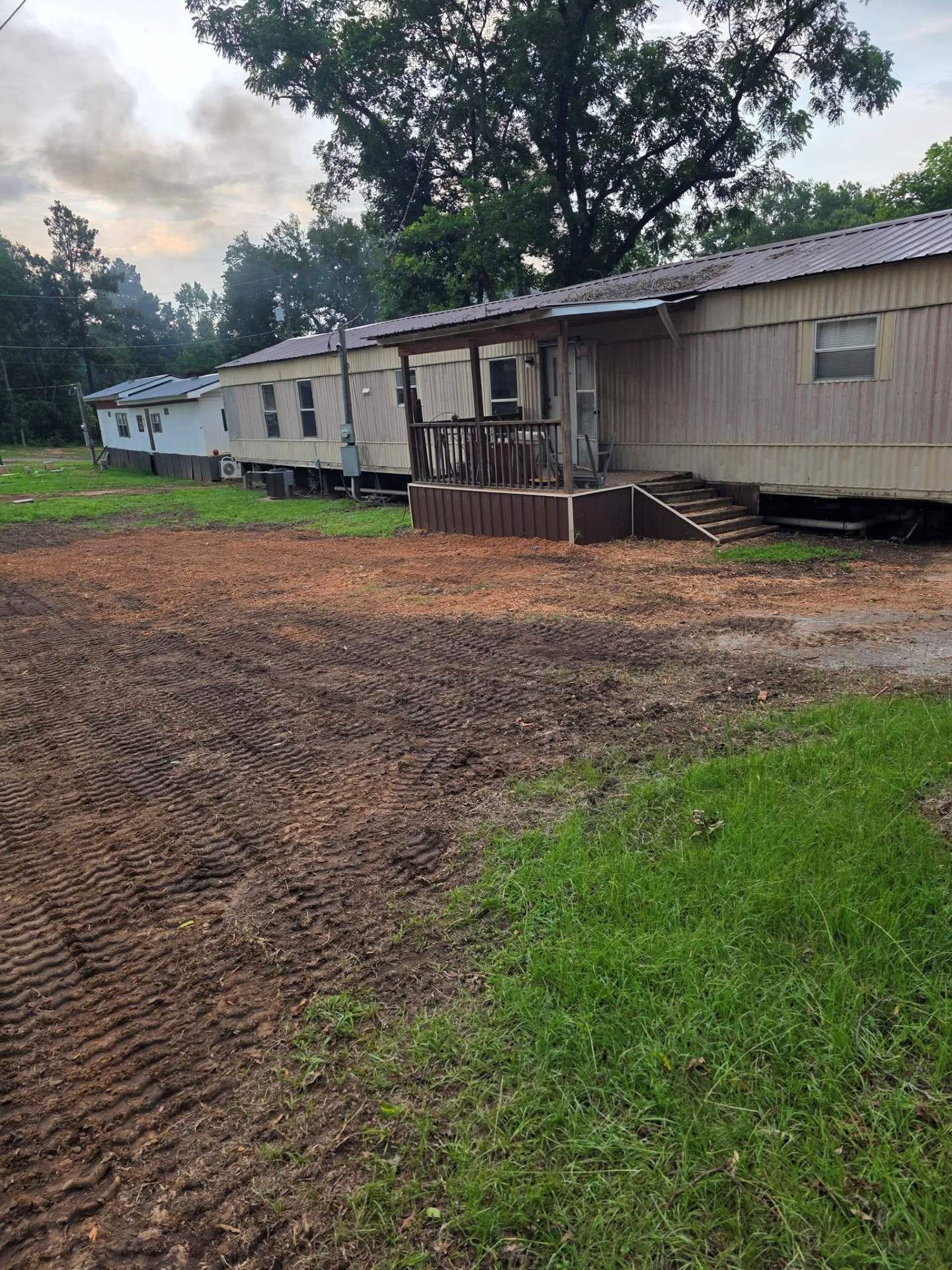 Long beige mobile home with a small porch and dirt yard under cloudy sky