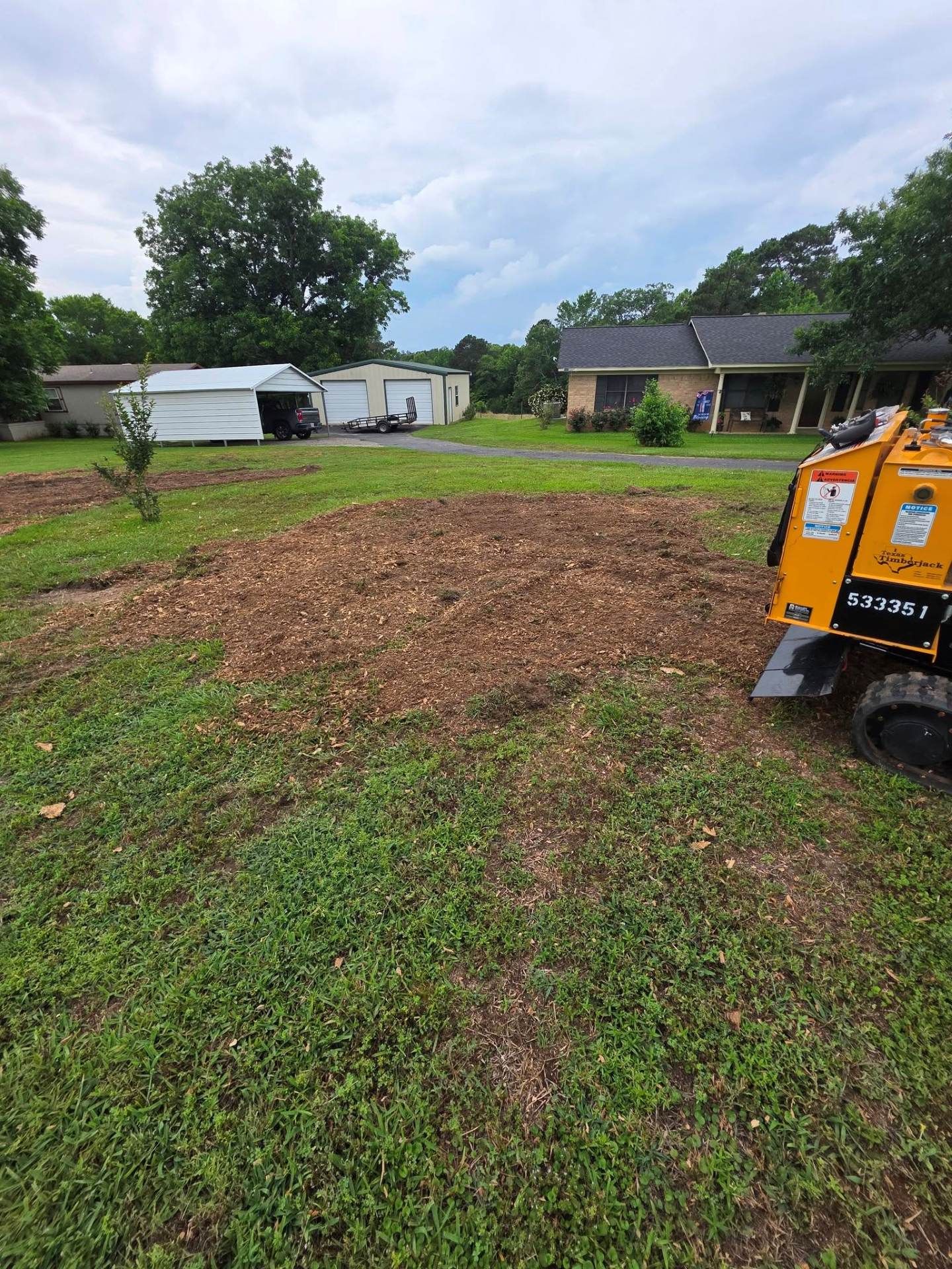 Muddy yard with a mulch pile in the center and an orange machine at the right edge, near houses and trees.