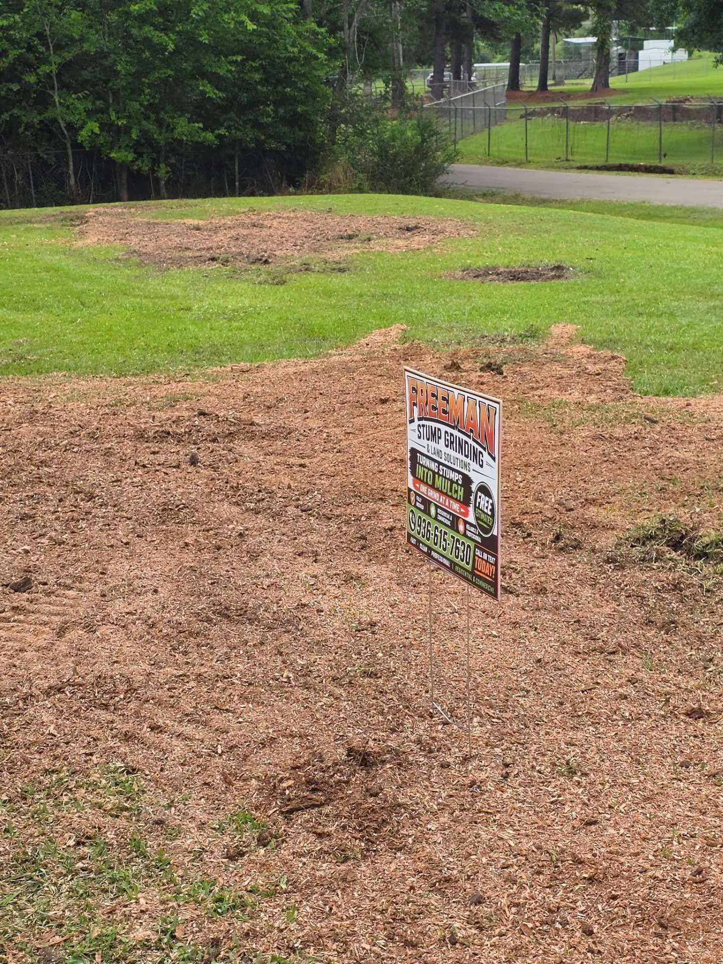 Wet dirt lot with a real estate sign in the foreground and a grassy park-like area behind it.