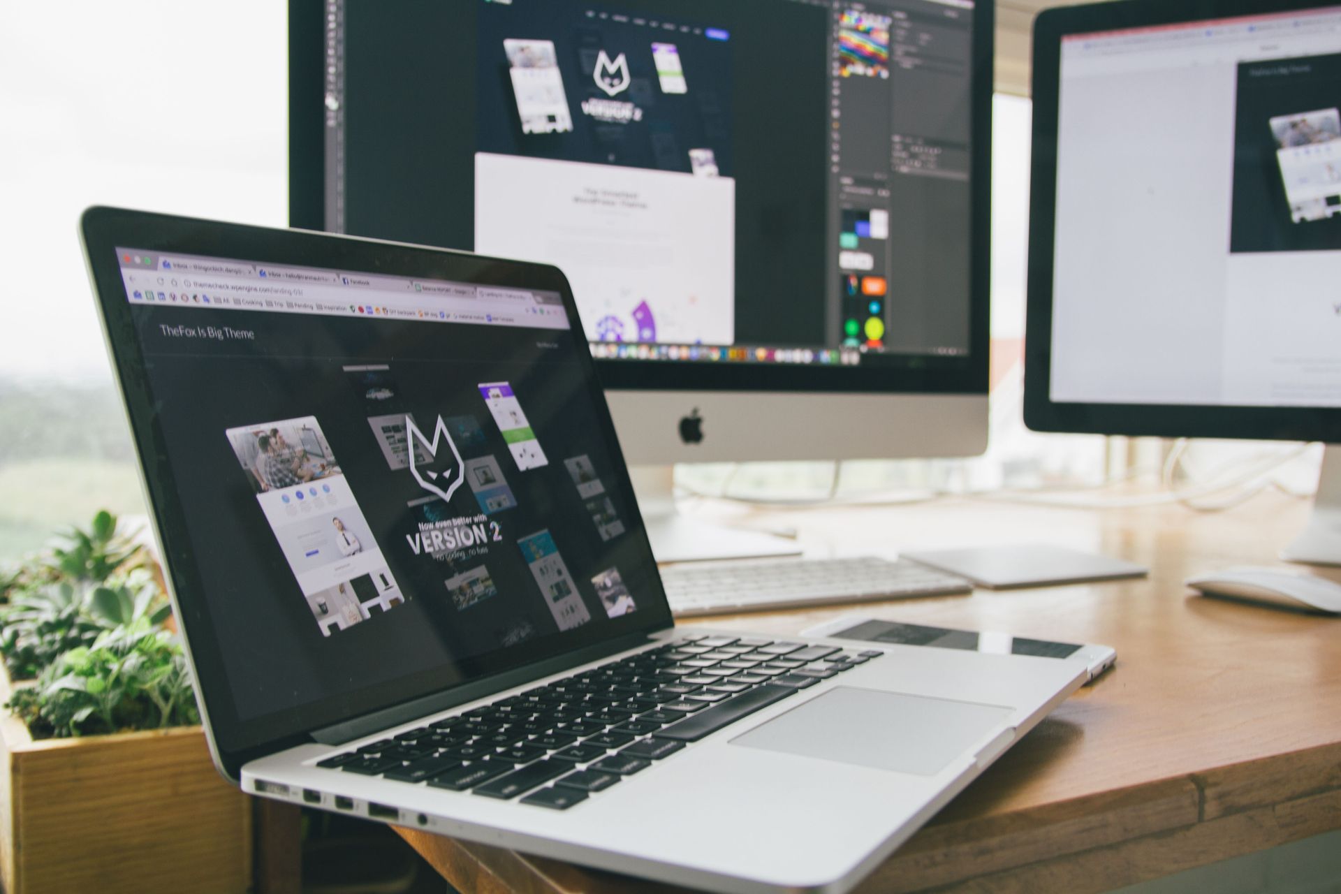 Laptop and desktop monitors on a wooden desk displaying design software in a bright office