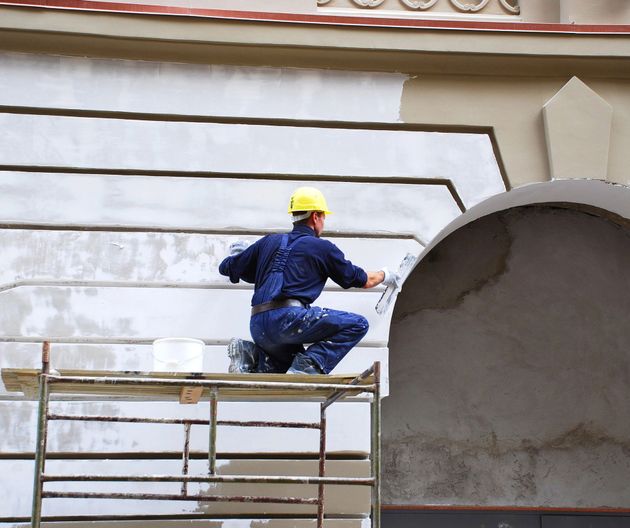 Un operaio con un elmetto giallo dipinge un muro esterno su un'impalcatura.
