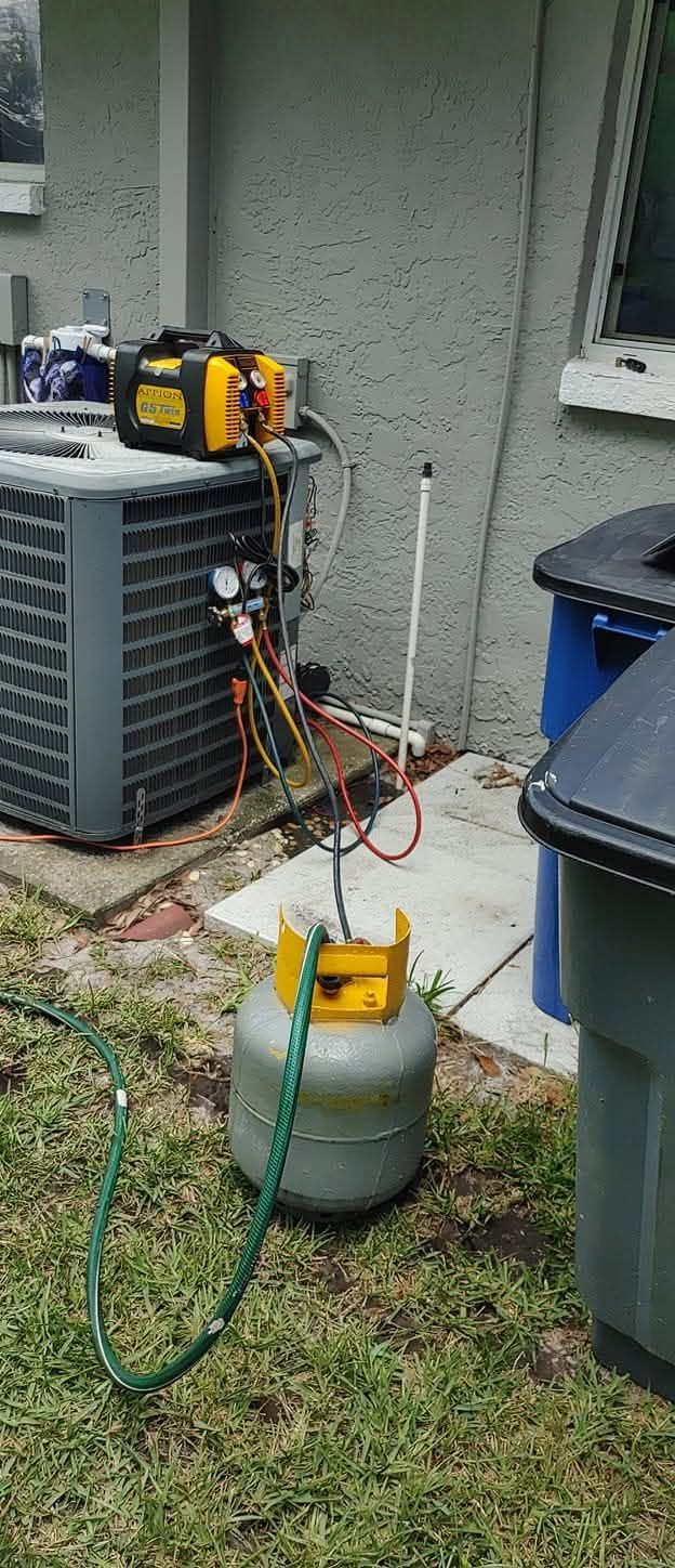 Outdoor HVAC units with gauges and hoses attached to a refrigerant tank on grass beside a house