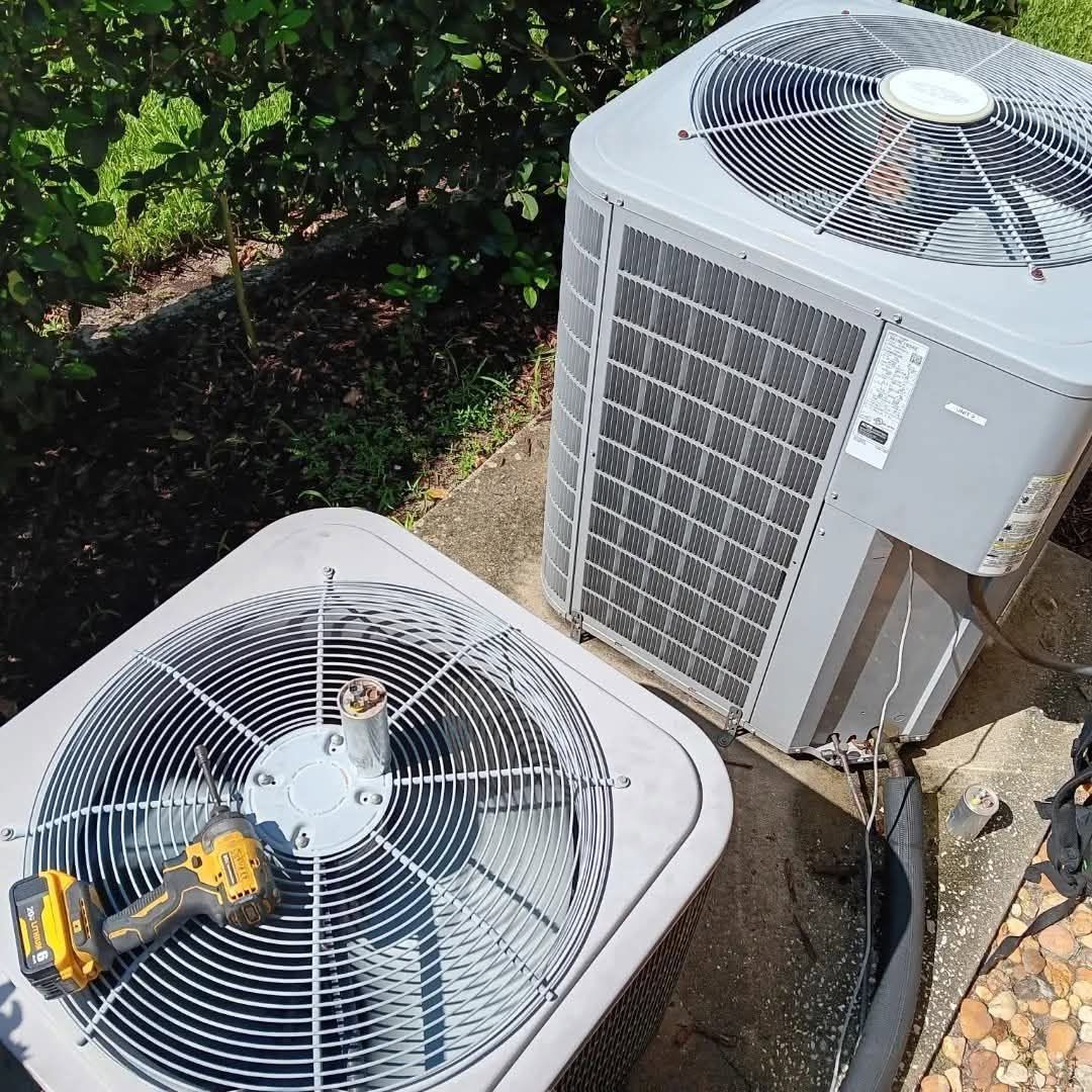 Two outdoor HVAC condenser units with visible fan grilles beside shrubs and a walkway