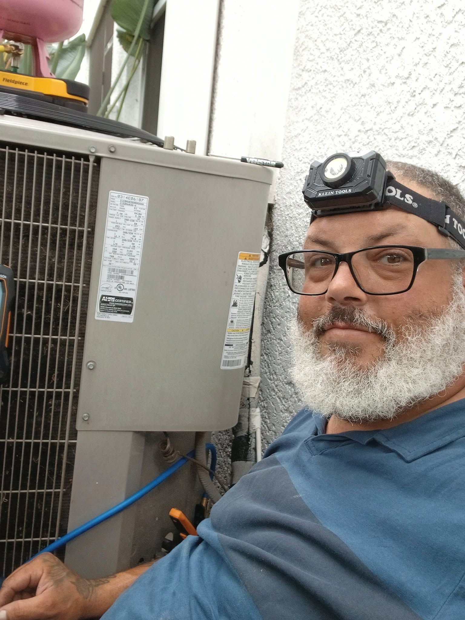 Technician wearing a headlamp beside outdoor electrical equipment, with a blue hose and fencing nearby.