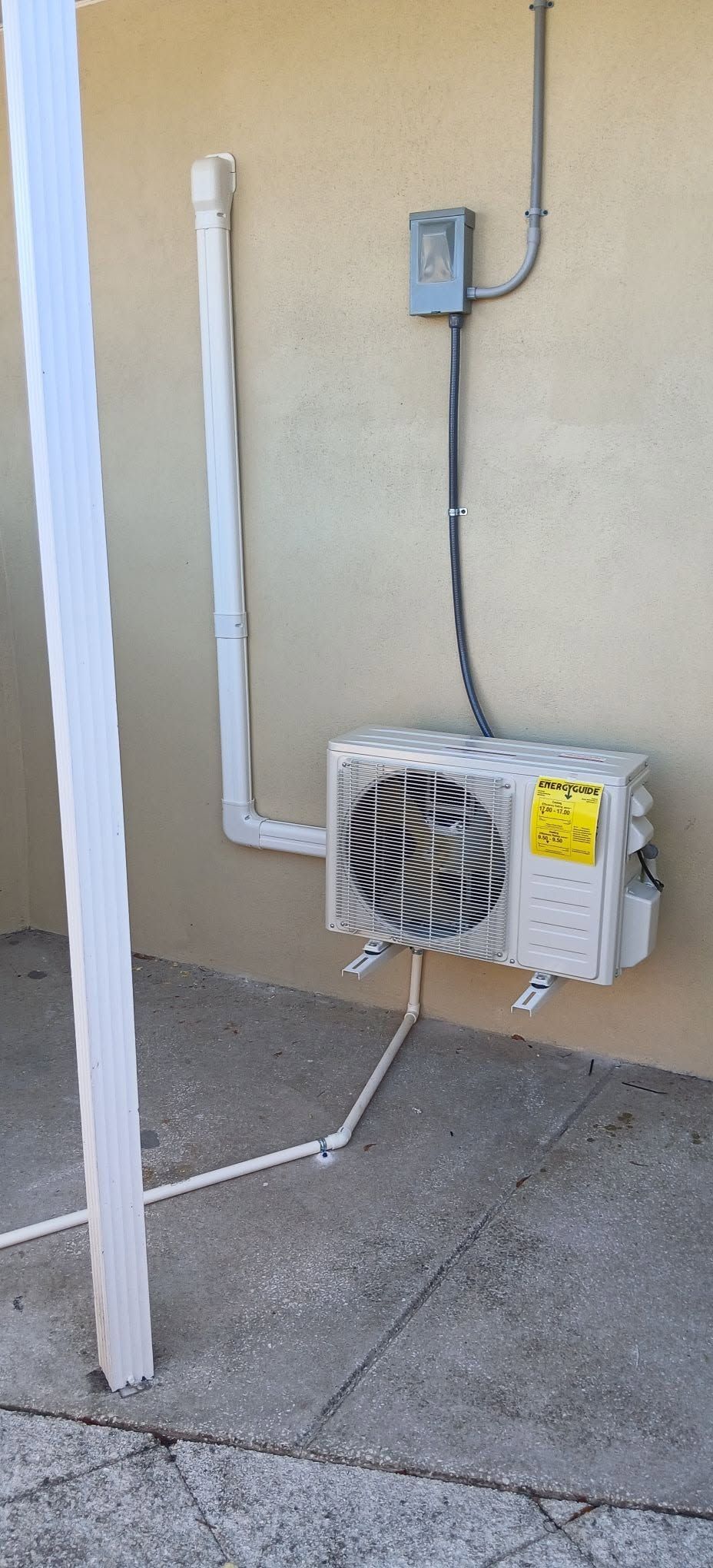 Outdoor HVAC unit mounted beside a wall with conduit and a nearby electrical box on a concrete pad
