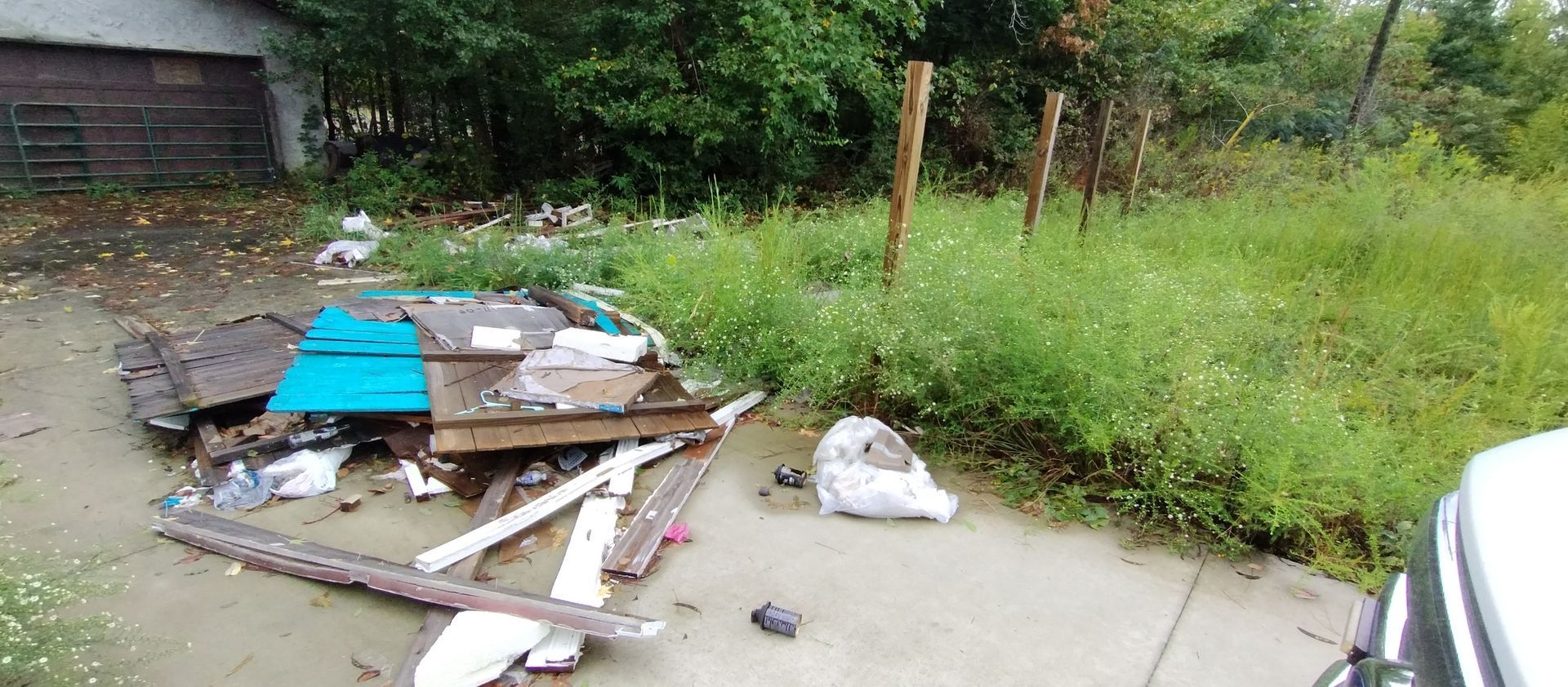 Debris and trash scattered on a paved driveway in front of overgrown greenery and a dilapidated garage. Debris and trash scattered on a paved driveway in front of overgrown greenery and a dilapidated garage.