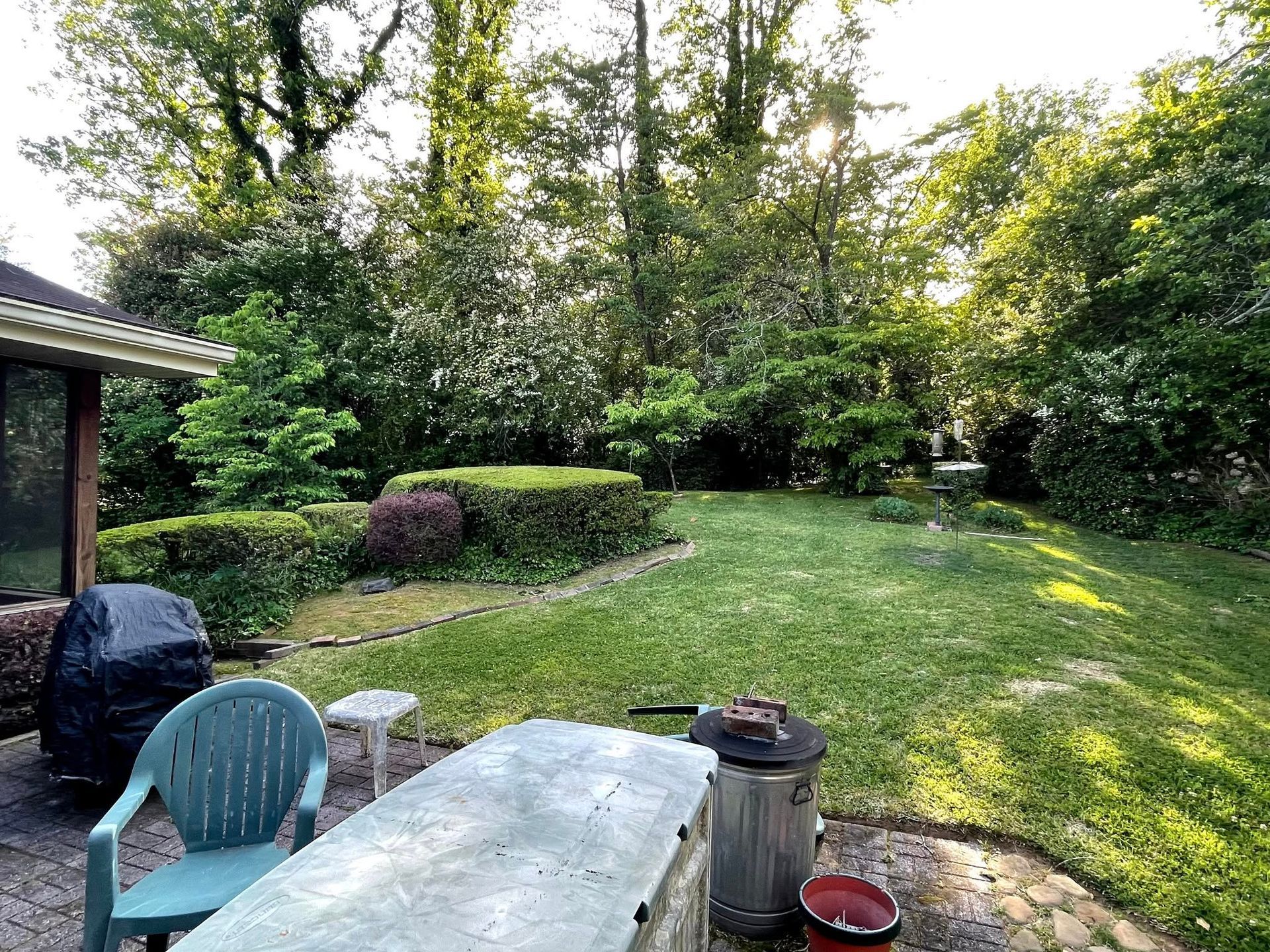 A sunny backyard with a stone patio, patio furniture, a grill, a metal trash can, and a grassy lawn framed by trees.