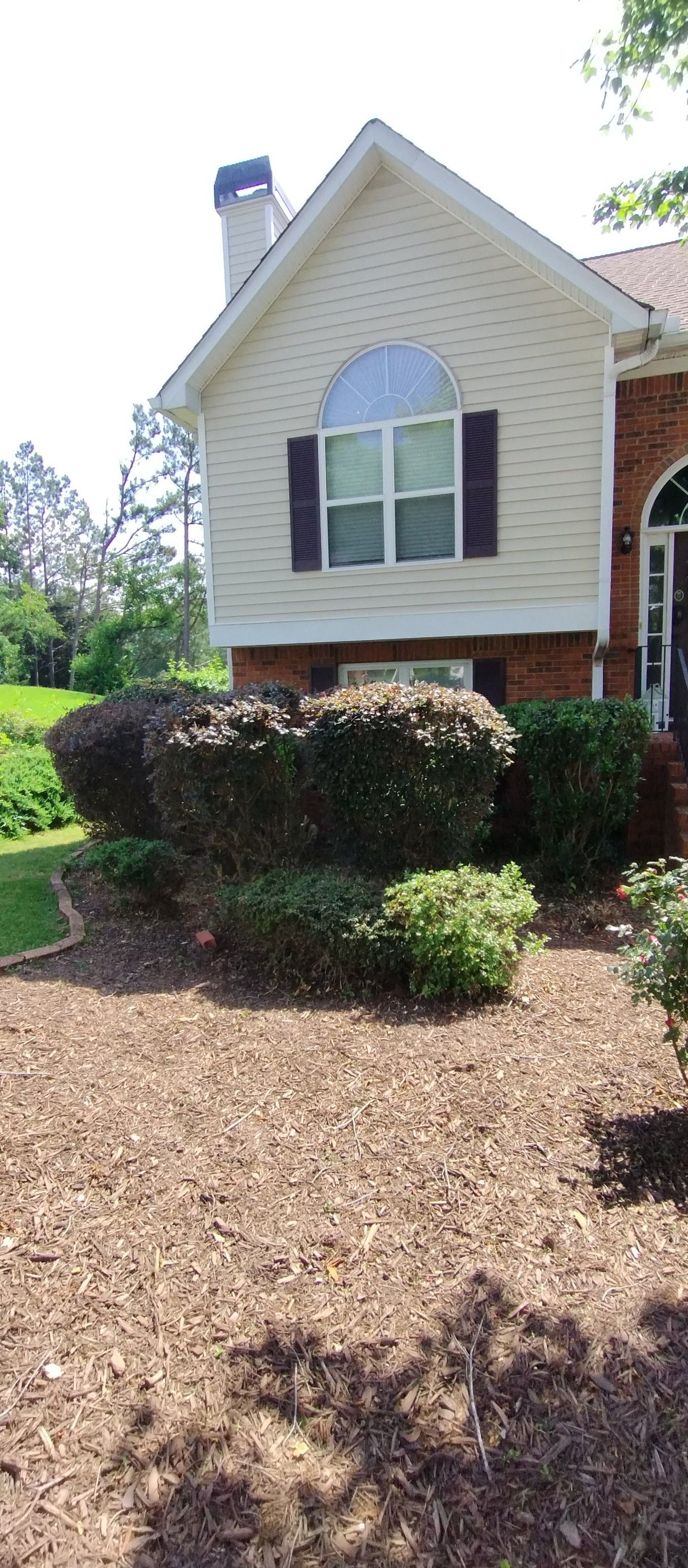 A suburban house exterior with light-colored siding, brick accents, arched windows, and manicured shrubs in the yard.