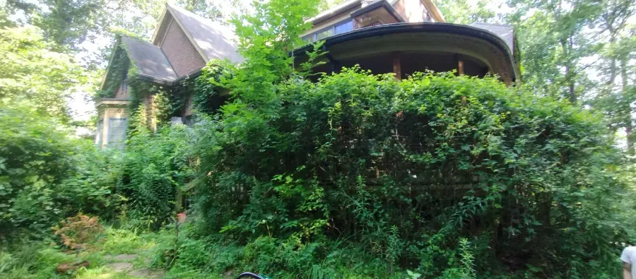 A two-story Victorian-style house largely obscured by dense green foliage and trees in a wooded setting.