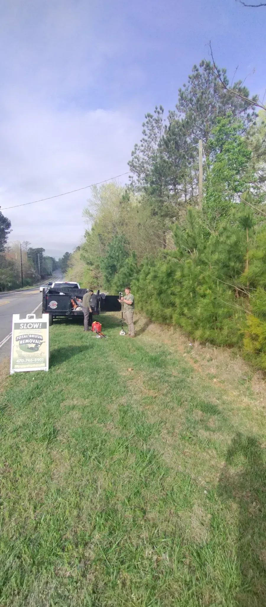 A person stands beside a parked dark vehicle on the grassy roadside near a white sign and a dense line of trees.
