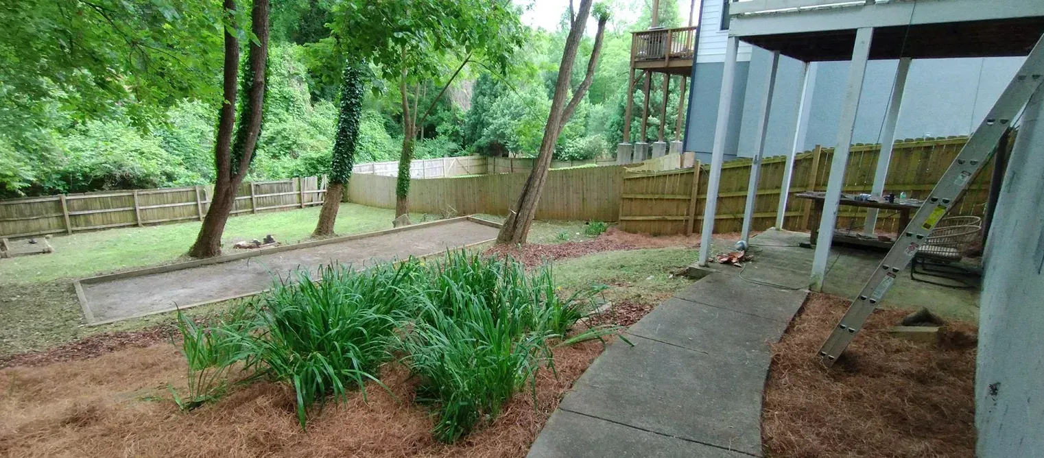 A backyard with a bocce ball court, a concrete path under a deck, a garden bed, and a wooden fence near dense trees.