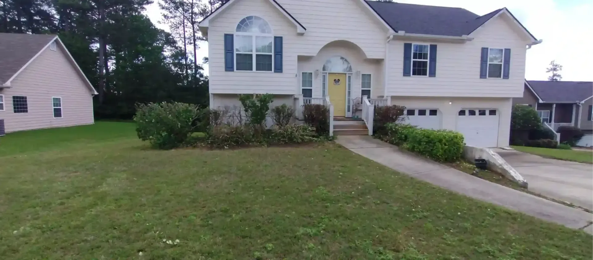 A light-colored two-story suburban house with a yellow front door, blue shutters, and a concrete driveway.