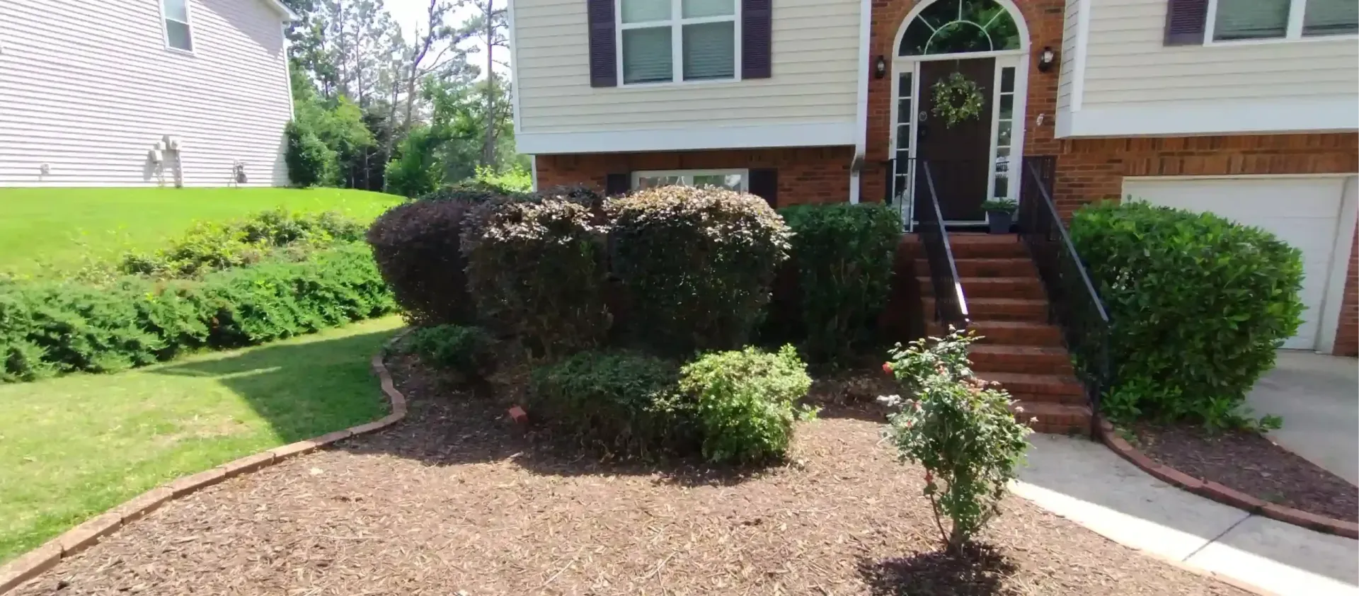 A front view of a two-story home with a brick-stepped entrance, landscaping shrubs, and a mulch-covered front yard.