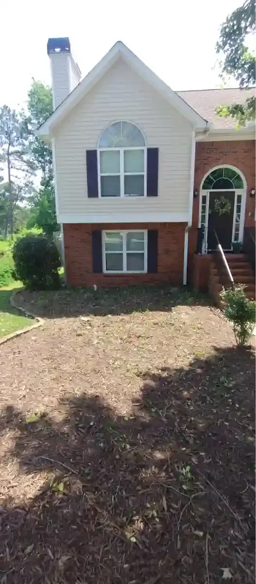 A two-story brick and white siding suburban house with a chimney and a mulched front garden bed under a bright sky.