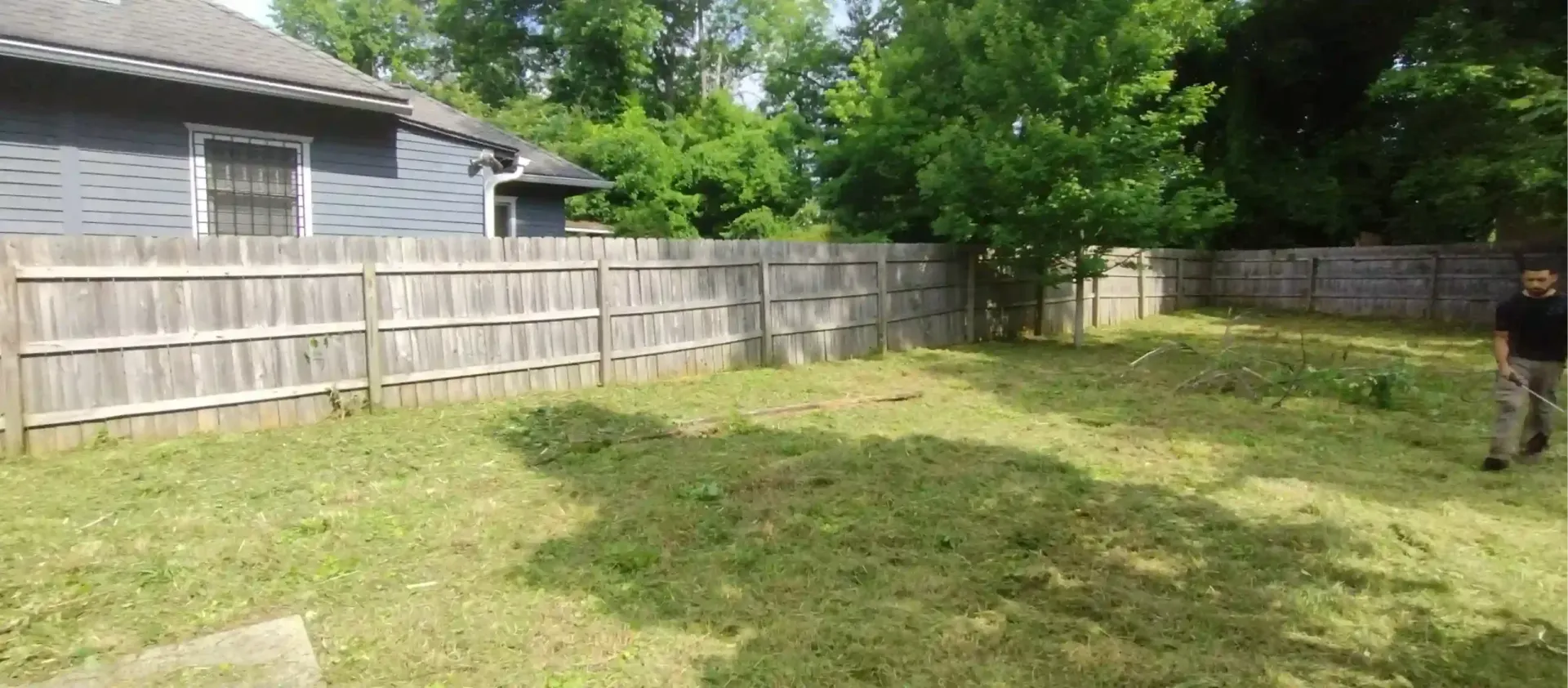 A person using a string trimmer to clear weeds in a backyard with a wooden fence and a blue house.