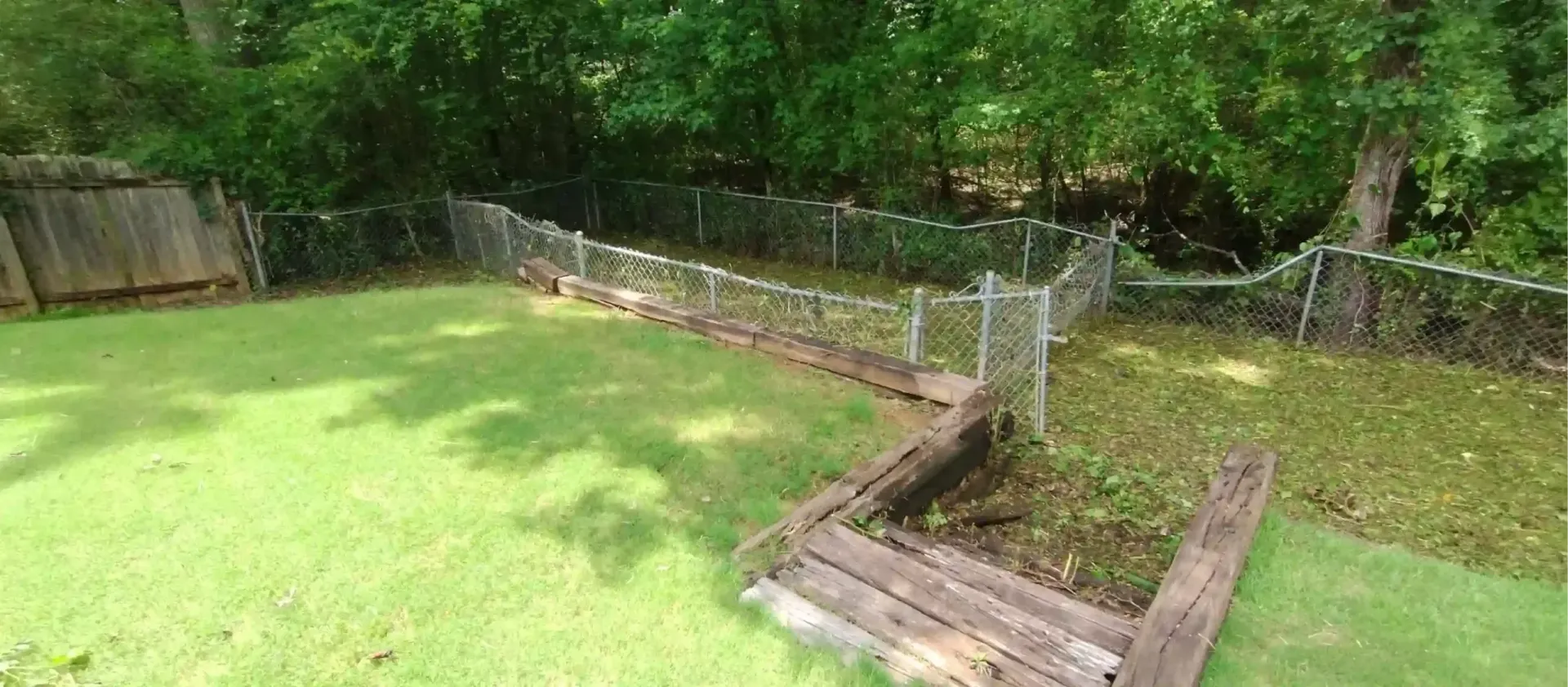 A grassy backyard with a wooden retaining wall, a small garden bed, and a chain-link fence bordering a wooded area.