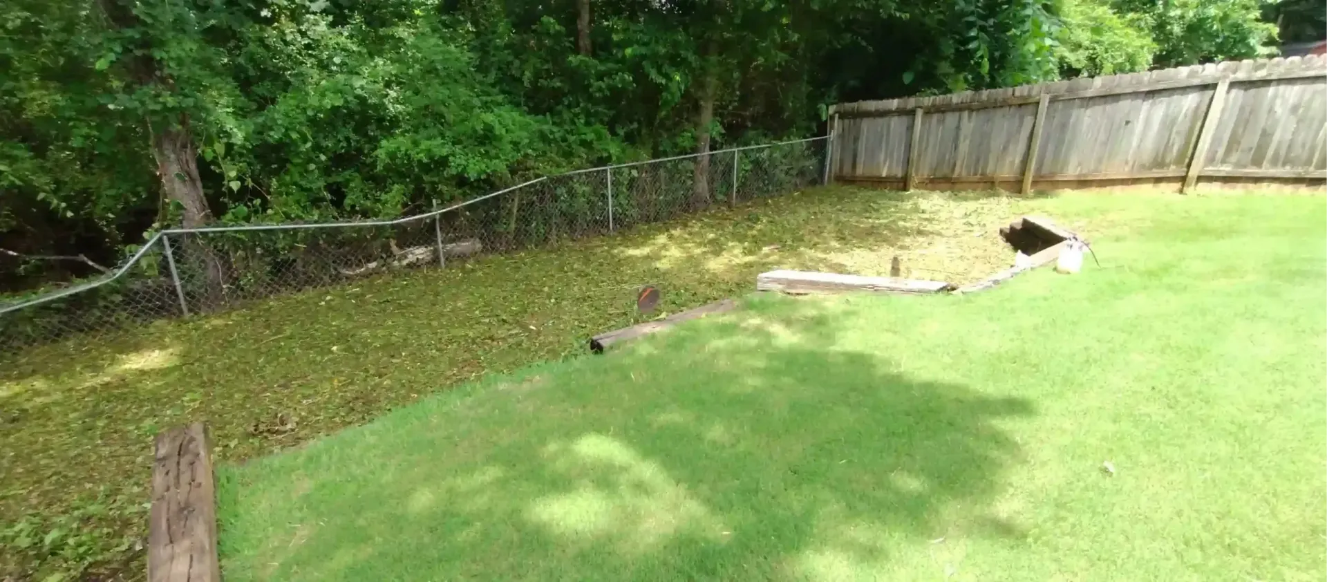 A backyard lawn features a mowed grass section, a patch of fallen leaves, a small wooden retaining wall, and a fence.