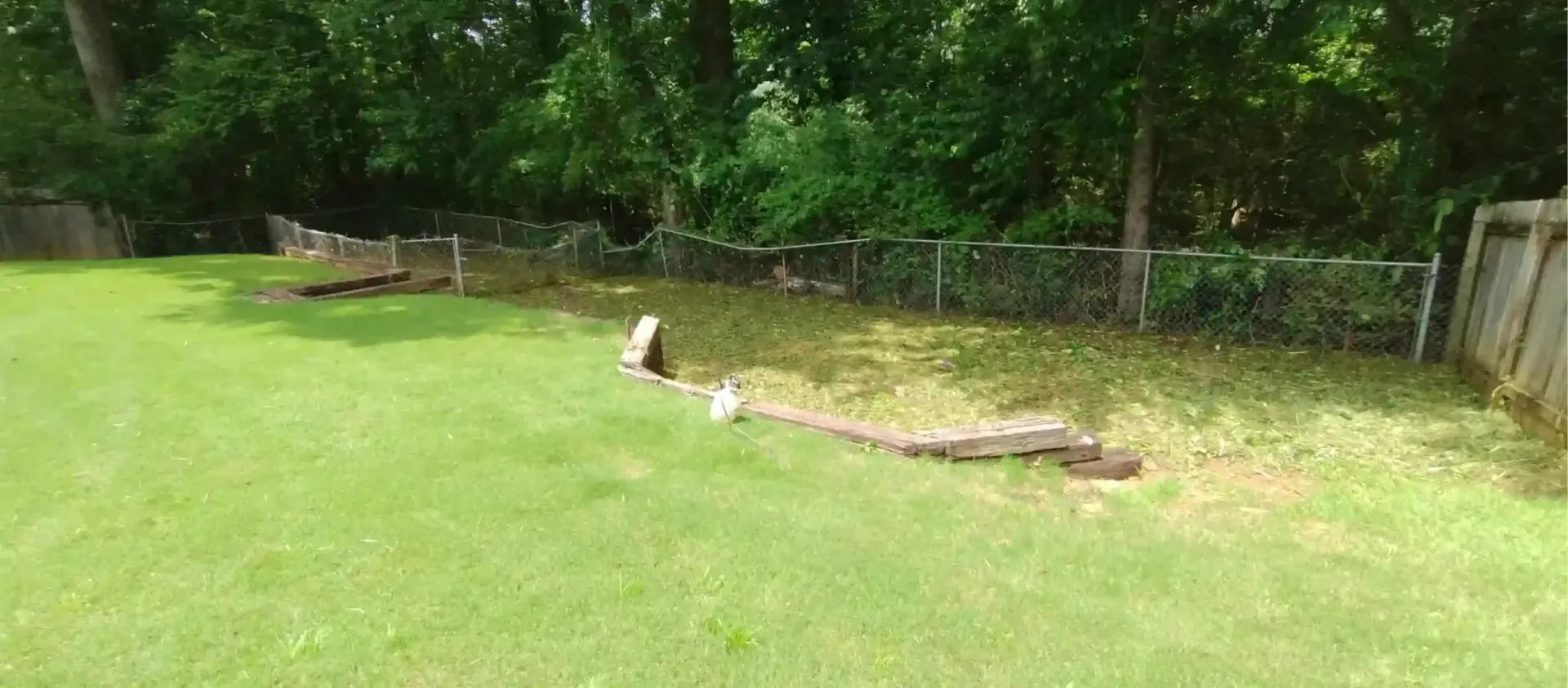 A grassy backyard with a low landscape timber retaining wall edging a mulched garden area near a chain-link fence.