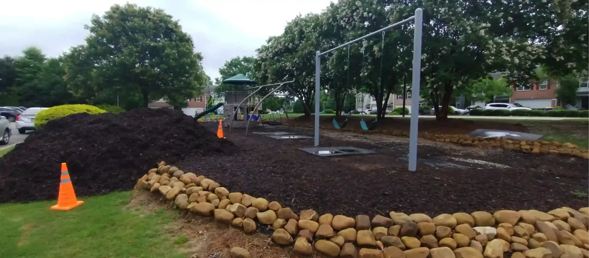 A playground with a swing set and slide, featuring a large pile of dark mulch and orange safety cones.