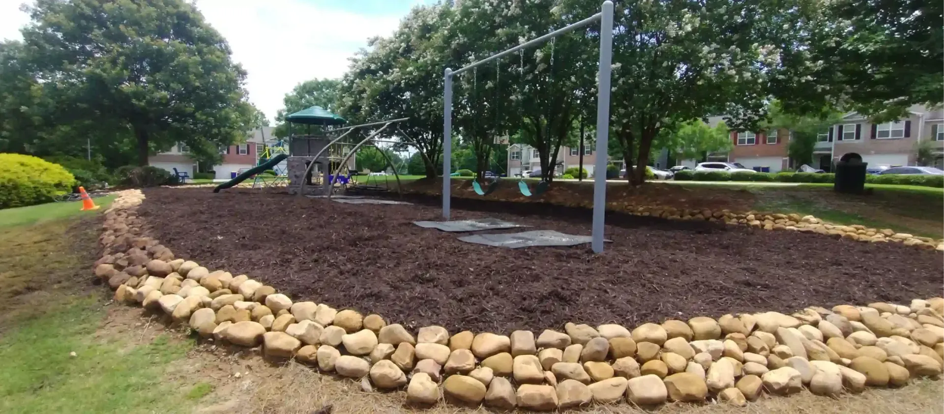 A small neighborhood playground with a swing set and a play structure, enclosed by a stone border and mulch ground cover.
