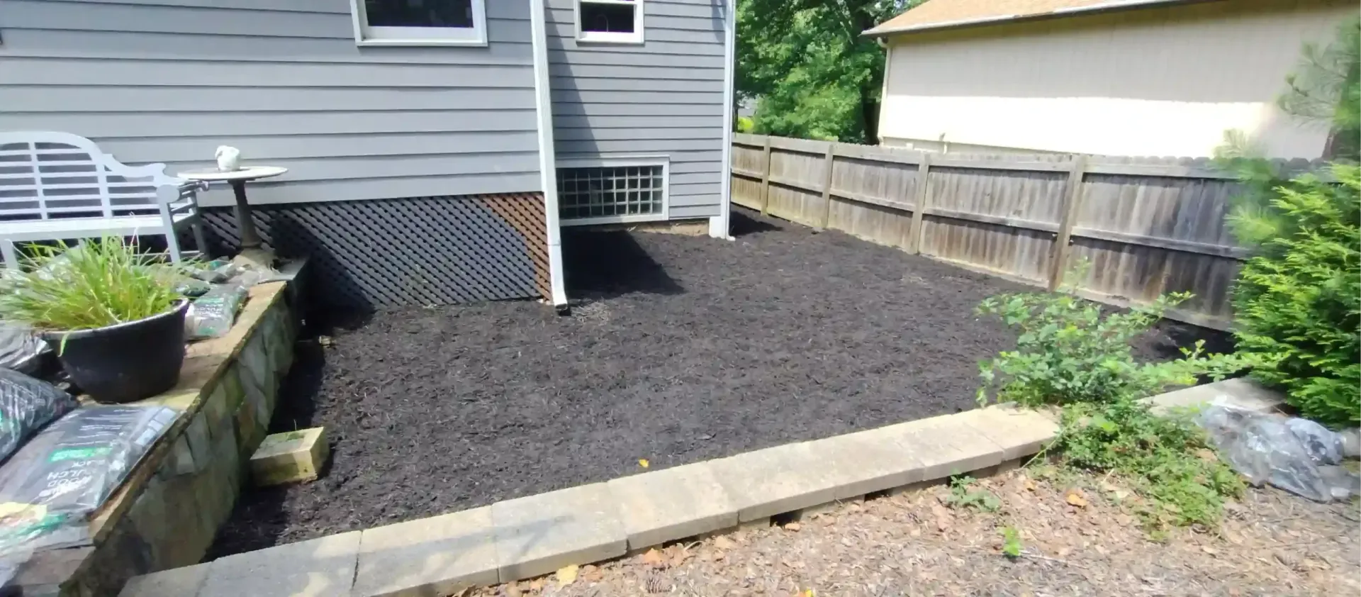 A patch of freshly laid dark mulch in a backyard garden area, bordered by a stone retaining wall and a wooden fence.