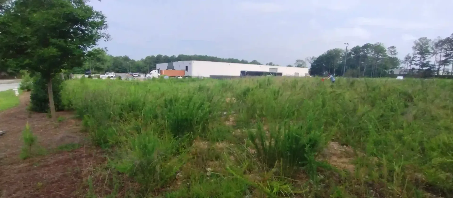 A large, grassy field in the foreground leads to a white industrial warehouse building under a cloudy sky.