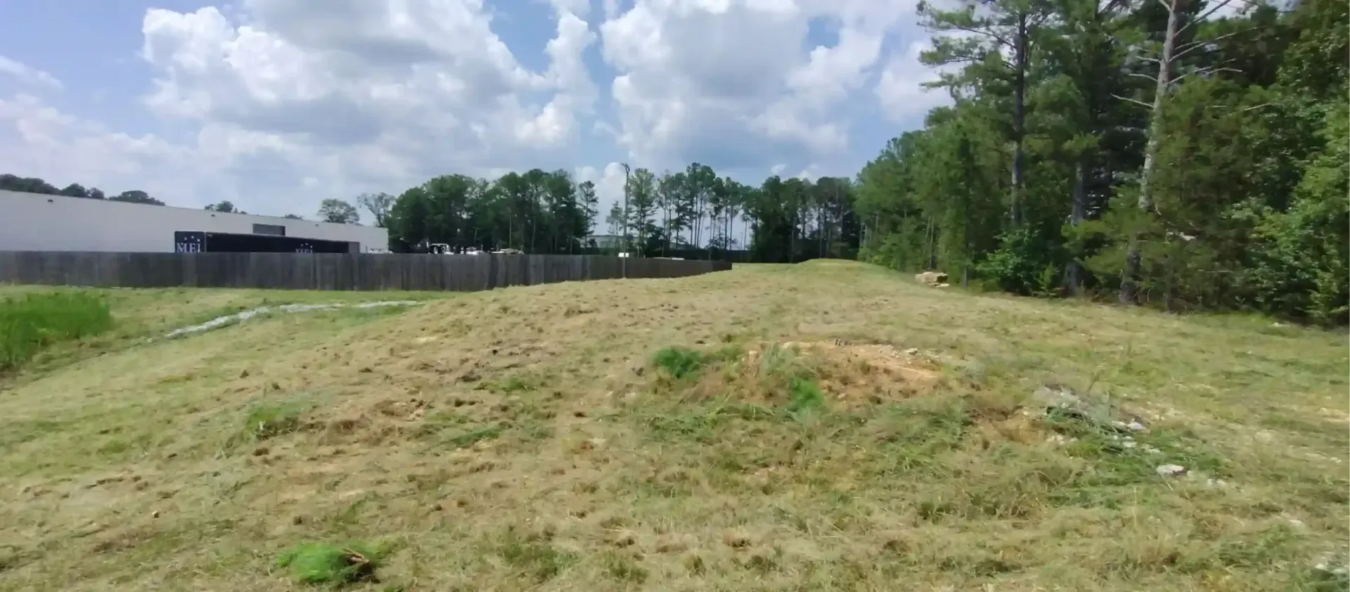 An open, grassy field sits adjacent to a fenced commercial building, bordered by trees on the right under a cloudy sky.