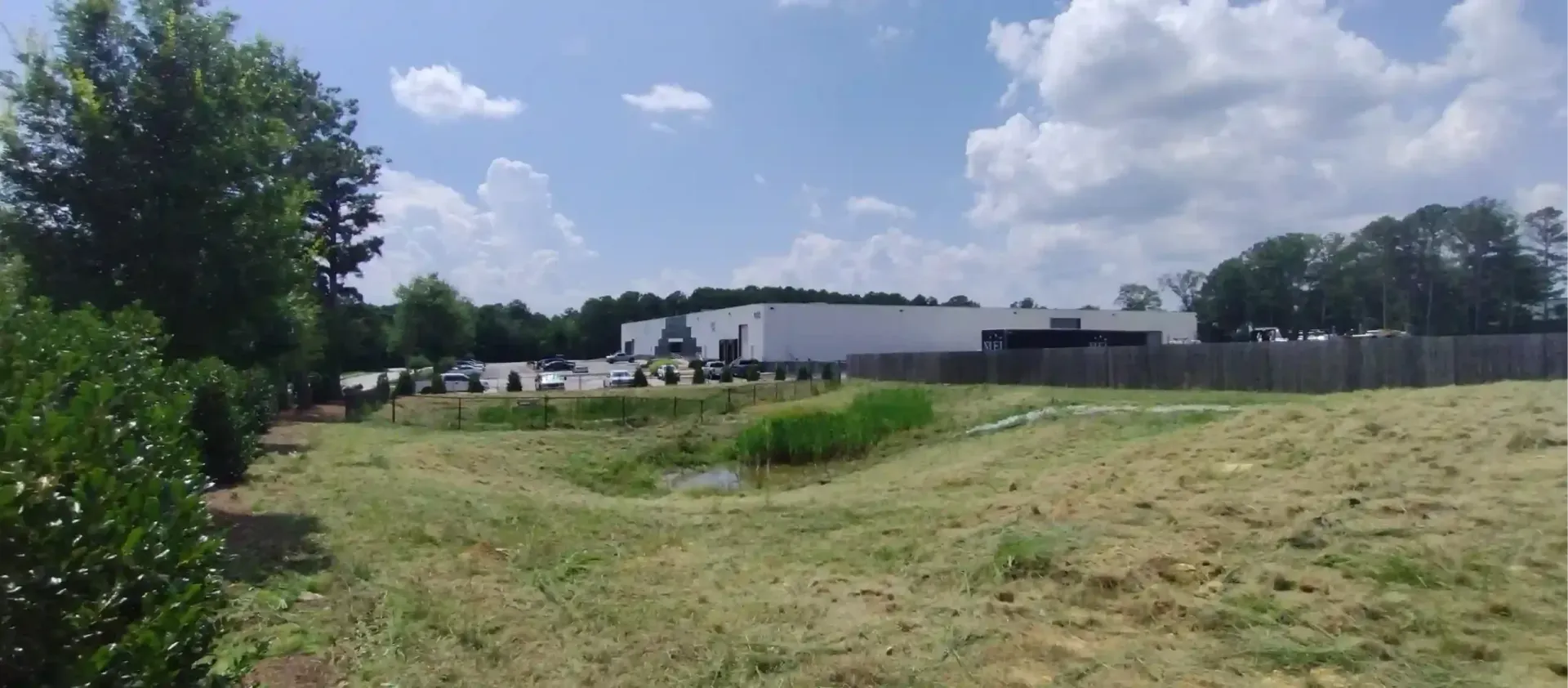 A large, white industrial building stands behind a grassy field and a small retention pond under a bright, cloudy sky.