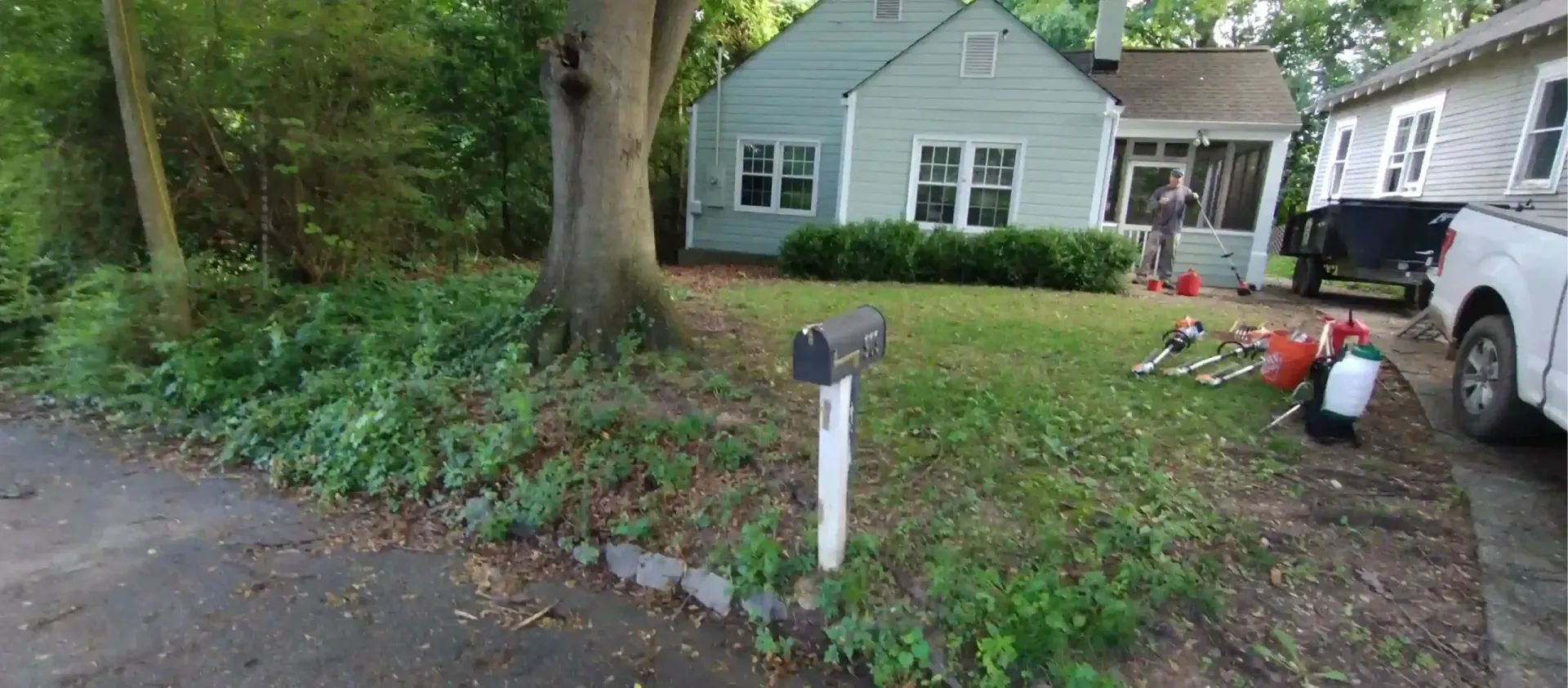 A light green, single-story house with a white front porch and a large tree in the front yard.