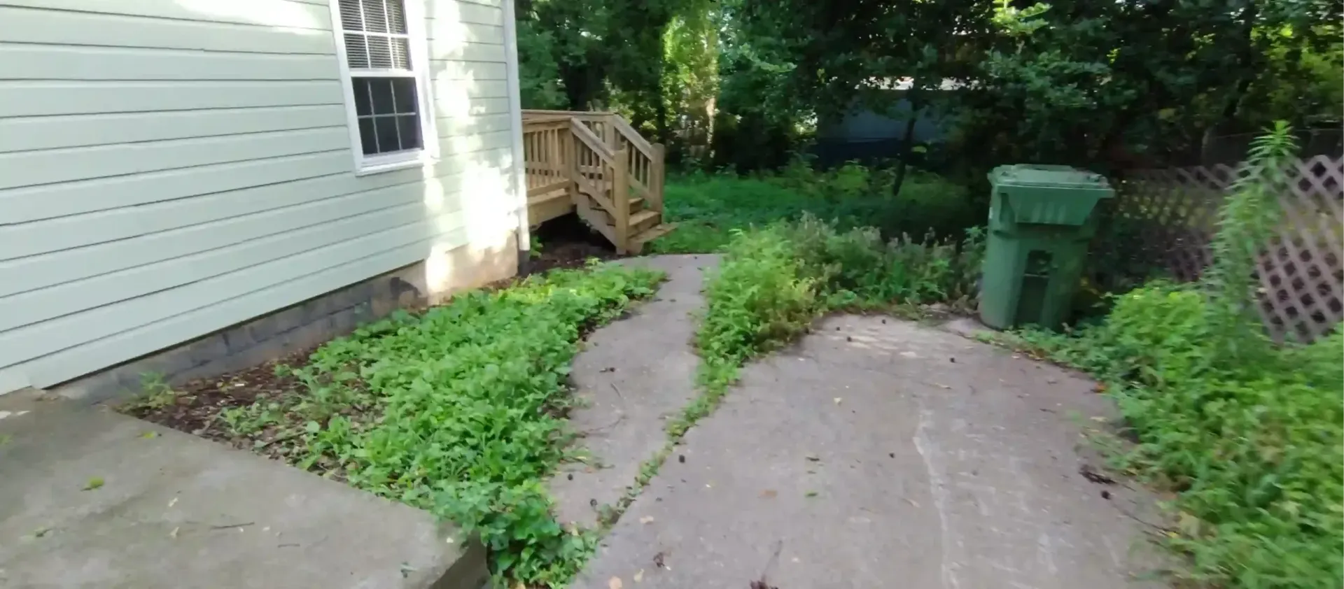 A concrete path winds through an overgrown backyard toward a small wooden deck next to a light-colored house.
