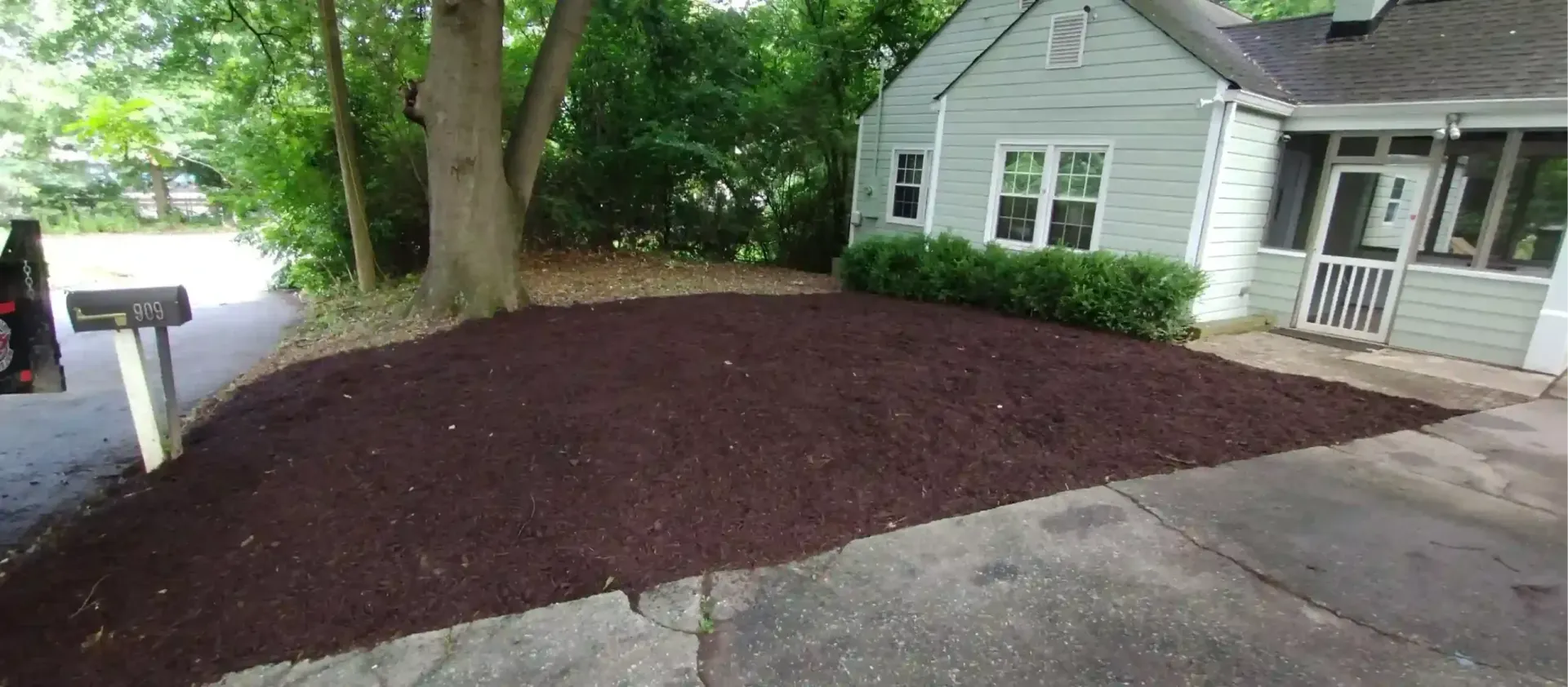 A fresh layer of dark brown mulch covers a large garden bed in front of a light-colored cottage-style house.