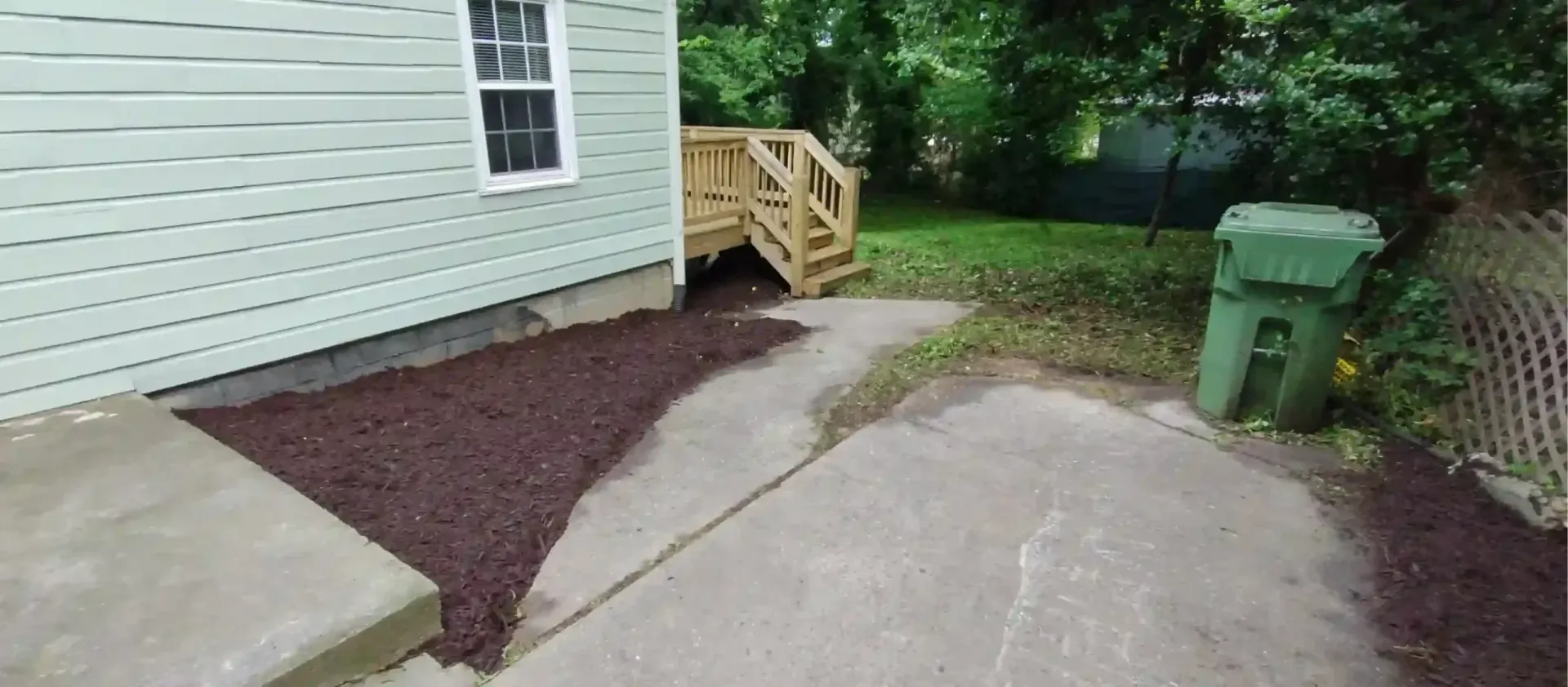 A light green house exterior with a wooden deck, a concrete patio, and a section of fresh dark brown mulch.