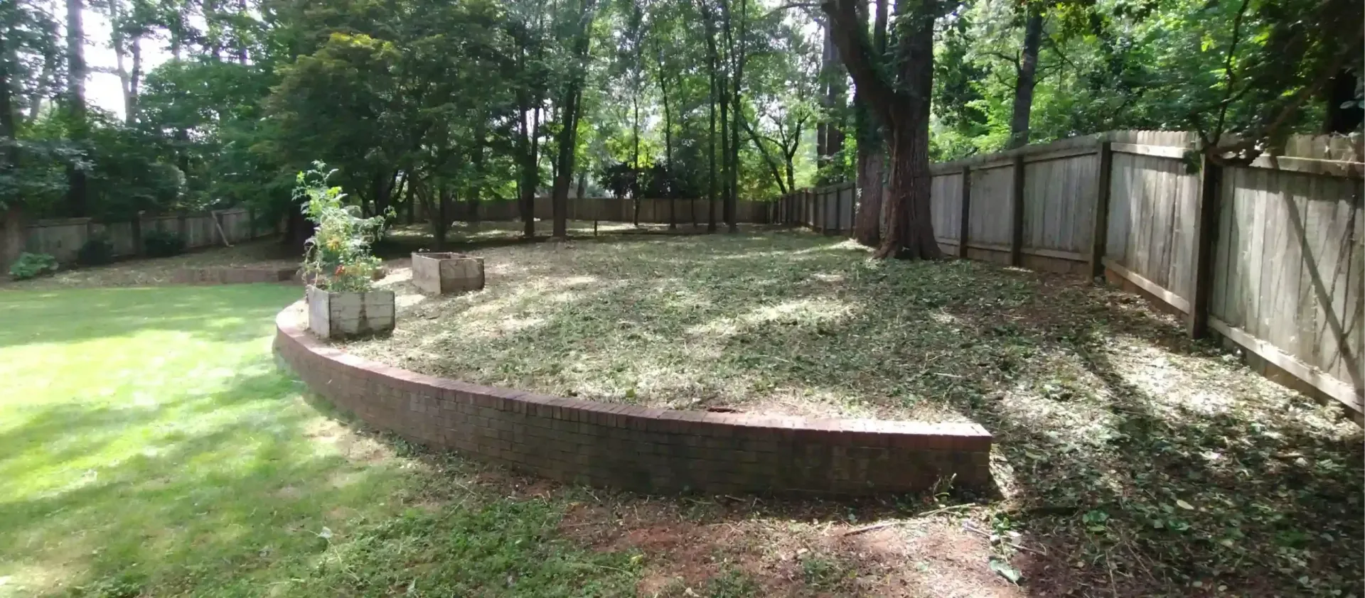 A curved brick retaining wall in a backyard with a large, shaded area of trees and a wooden fence along the perimeter.