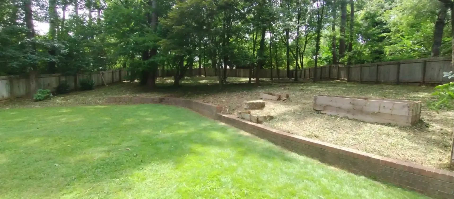 A sloped backyard featuring a manicured lawn, a low concrete retaining wall, and a raised planter box near a tree line.