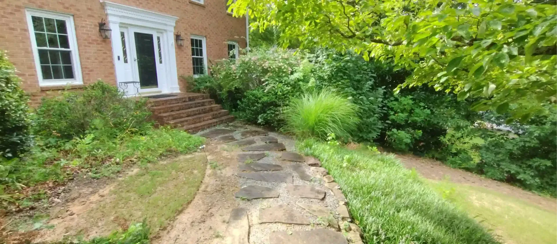 A stone walkway leads to the front entrance of a brick house surrounded by green trees and shrubs.