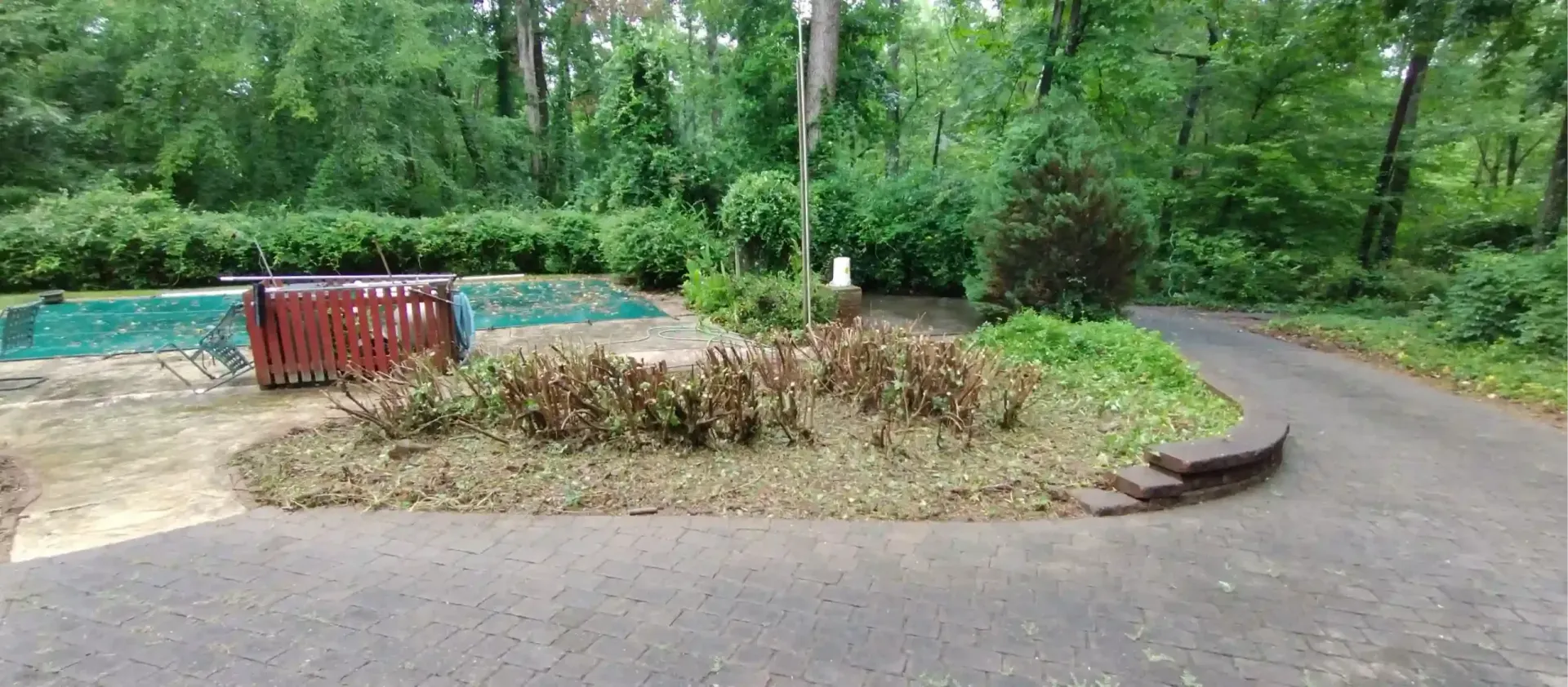 A paved driveway curves past a central landscaping island with dried shrubs, leading to a pool area near dense woods.
