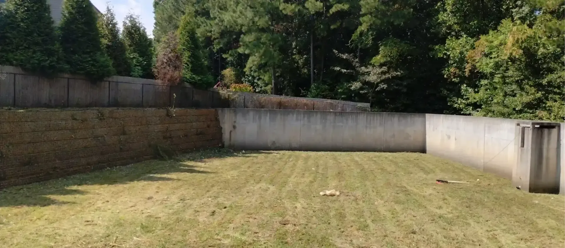 A grassy backyard enclosed by a stone retaining wall on the left and a smooth concrete wall on the right.