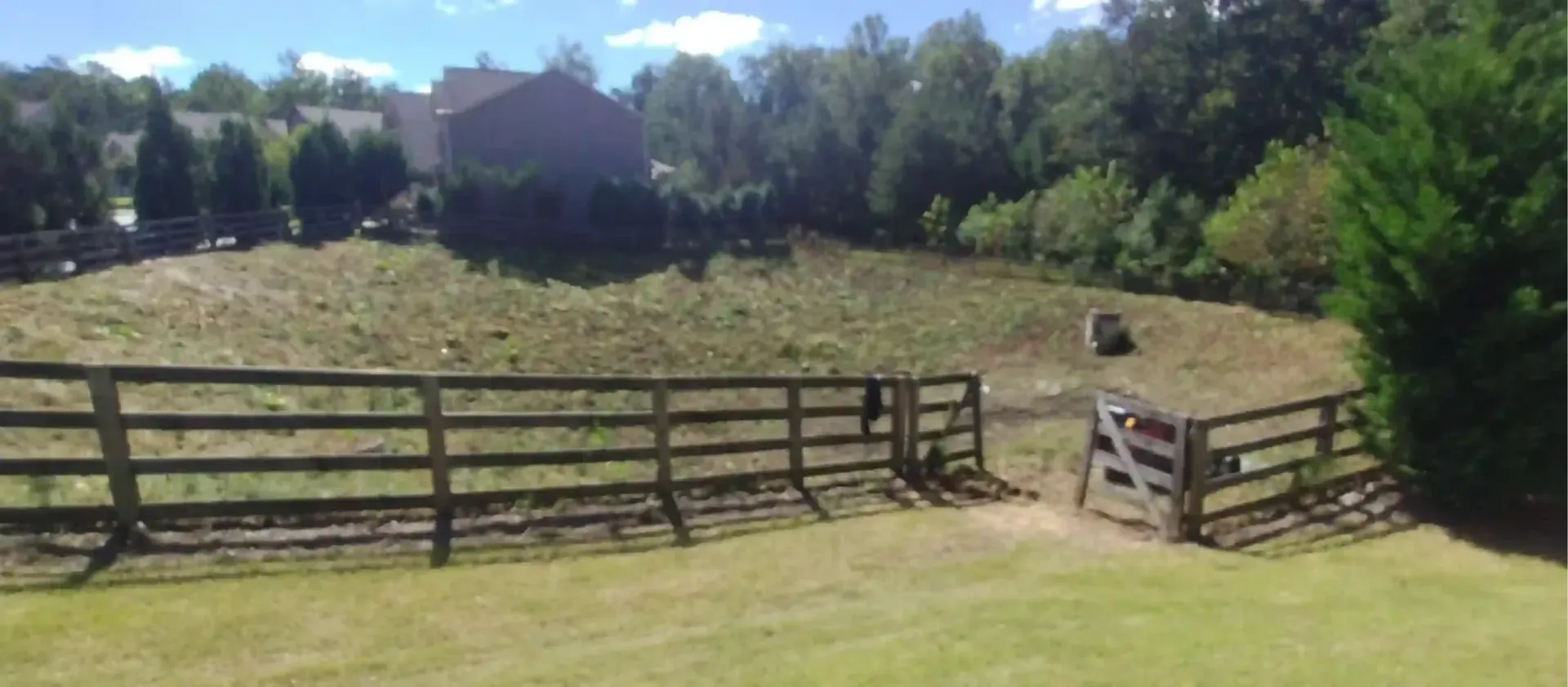 A grassy, sloping fenced pasture on a sunny day with trees in the background and a wooden gate on the right.