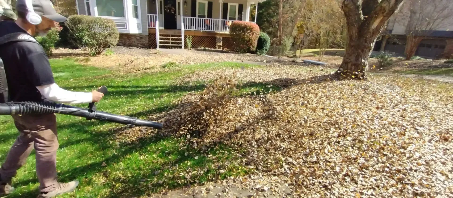 A person wearing headphones uses a leaf blower to clear brown, fallen leaves from a residential lawn.
