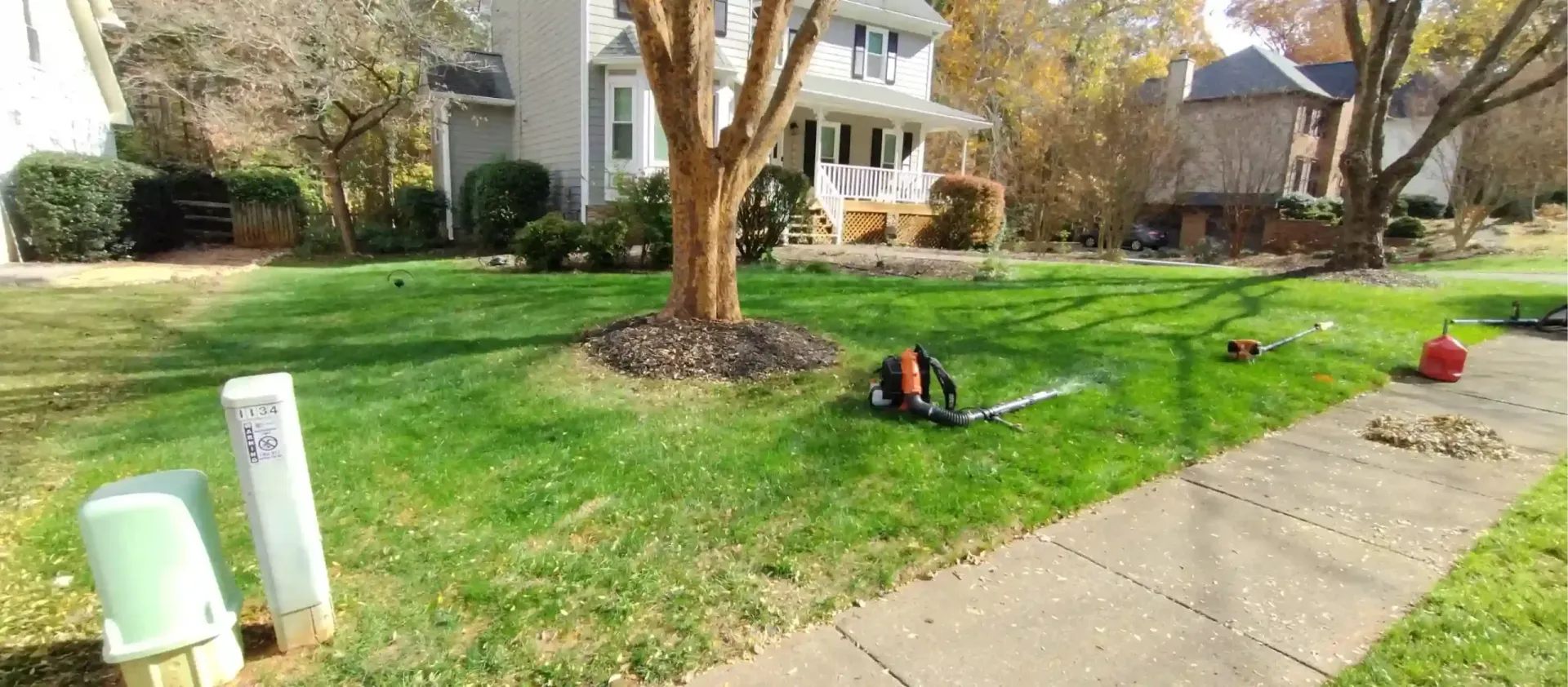 A residential front yard with a large tree in the center, a sidewalk, utility boxes, and lawn equipment scattered about.