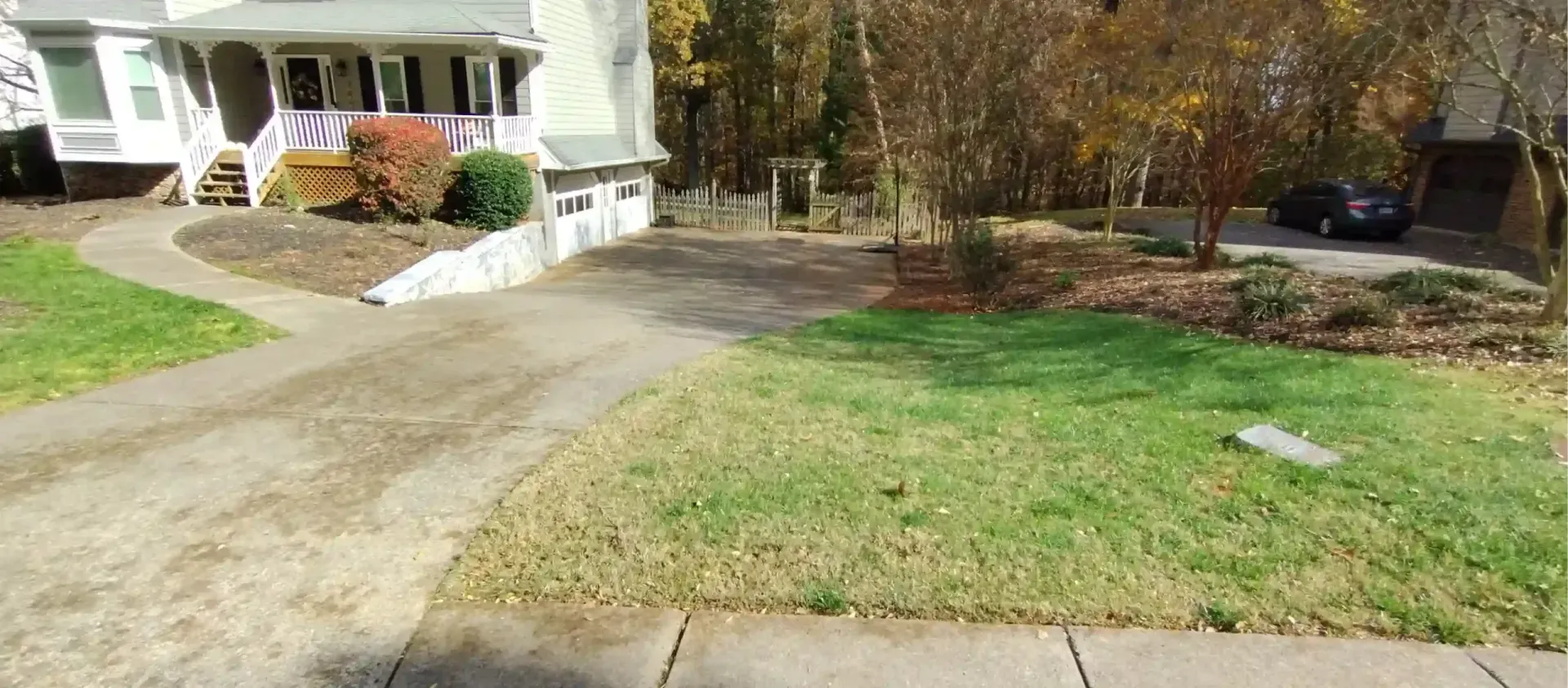 A driveway leads toward a two-story house with autumn trees, a grassy lawn, and a parked car in the background.