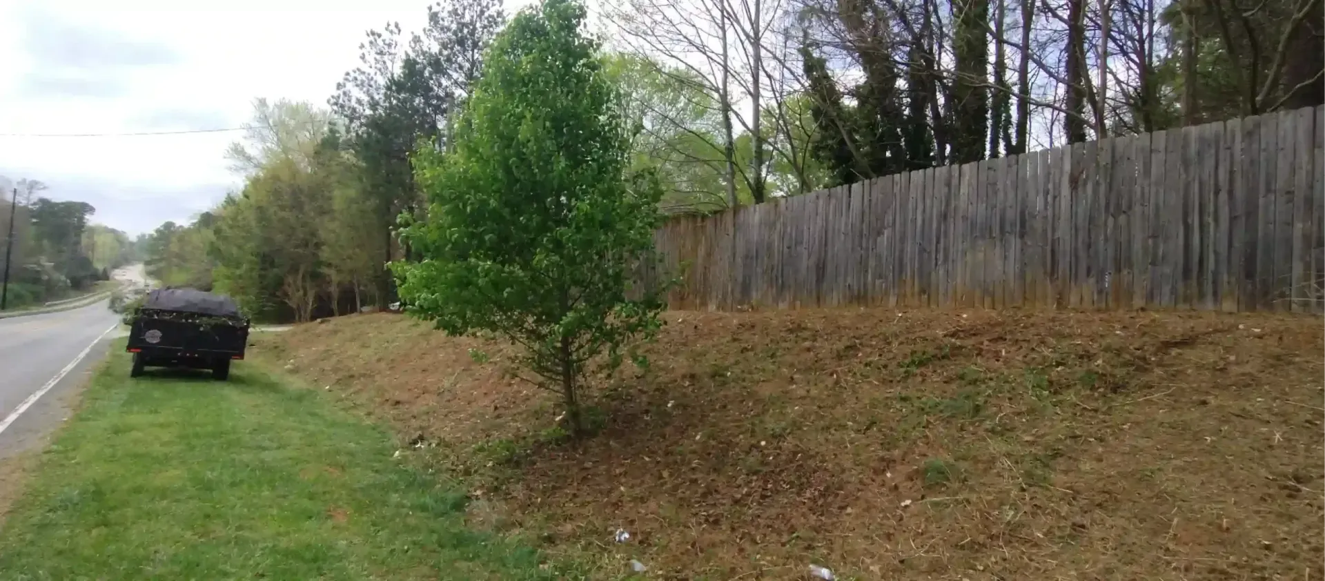 A dark vehicle is parked on a grassy shoulder next to a sloped, grassy area with a small tree and a tall wooden fence.